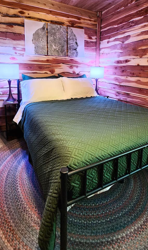 This cozy bedroom features warm cedar walls, a braided rug, and a plush queen bed with a deep green quilt. Soft lighting and a striking tree-ring wall art create a calming, nature-inspired space perfect for unwinding after a day outdoors.