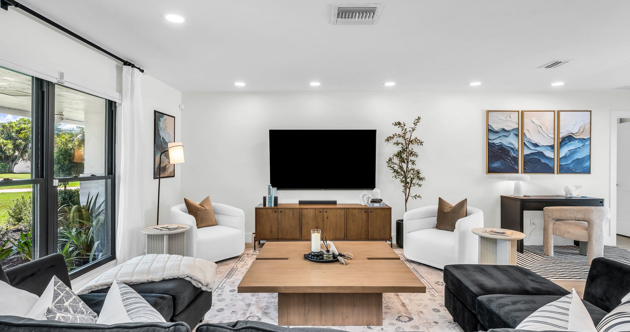 Elegant open-concept living room featuring a plush black velvet sectional, 75” smart TV, and designer mid-century modern furnishings. Floor-to-ceiling windows and French doors fill the space with natural light and connect seamlessly to the outdoor pool.