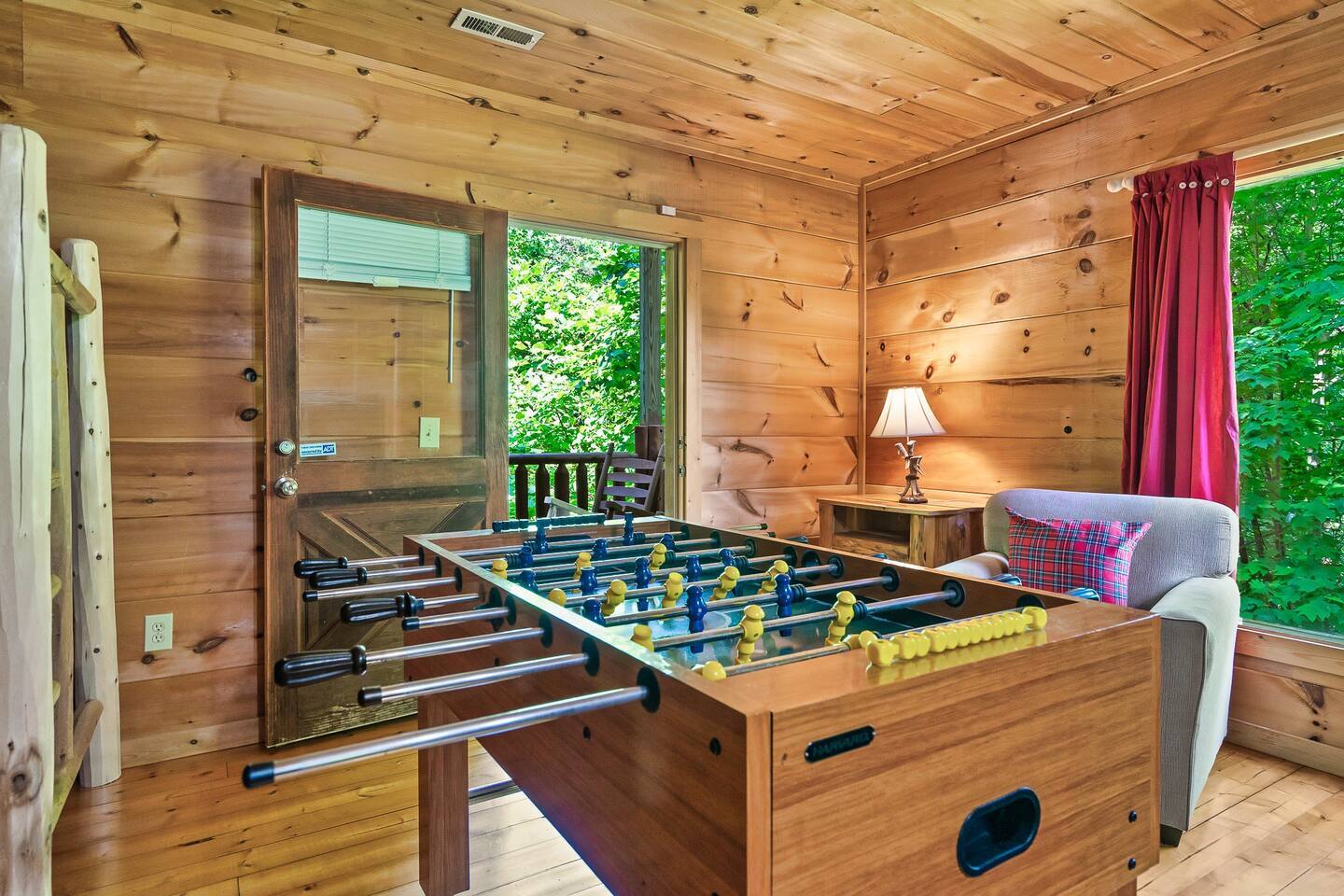 A charming game nook with a foosball table, soft lighting from a lamp, and an open door leading to lush green views, creating a lively yet peaceful atmosphere.