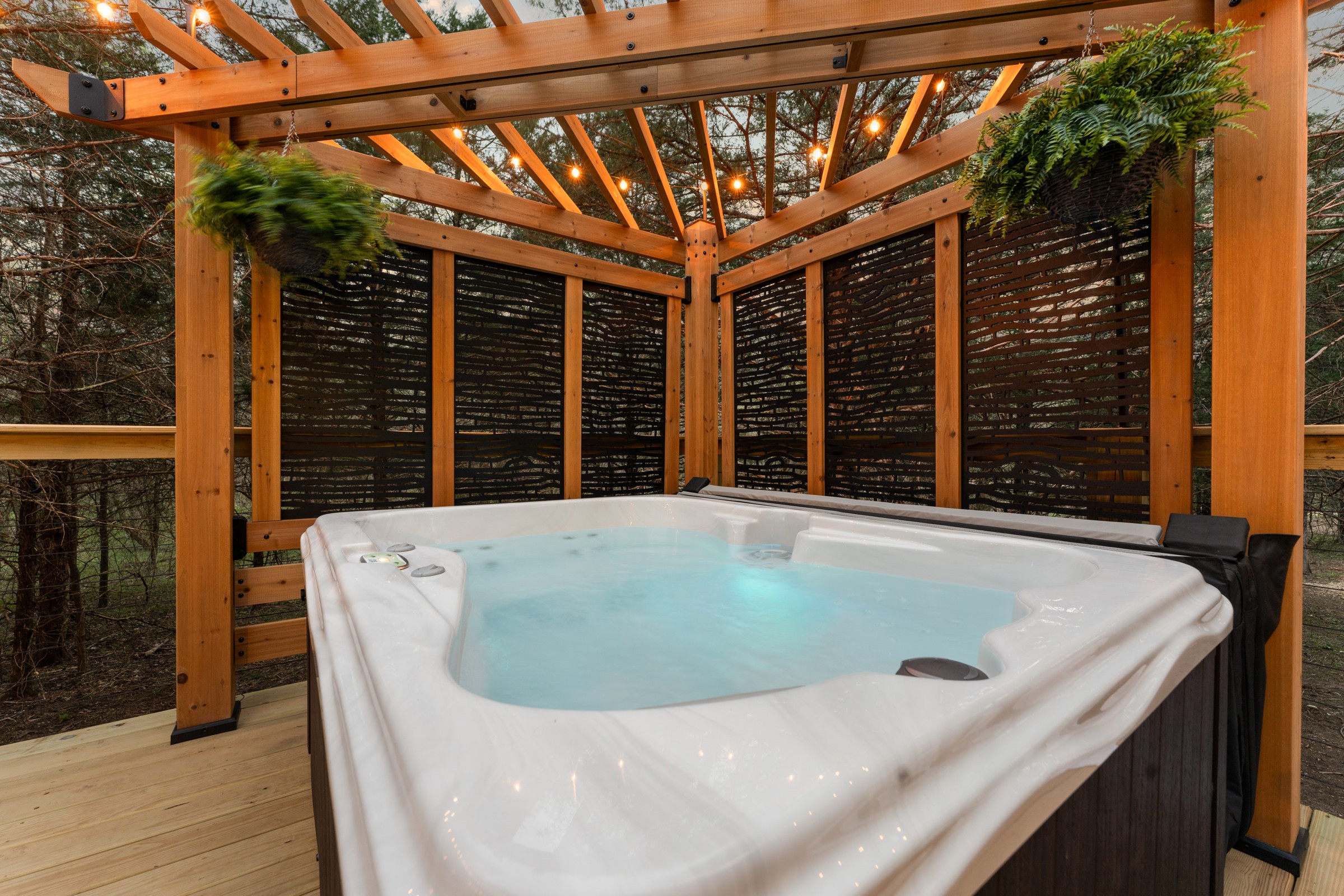 Hot tub under the pergola with hanging ferns. Privacy screens and string lights set the mood as the sun goes down.