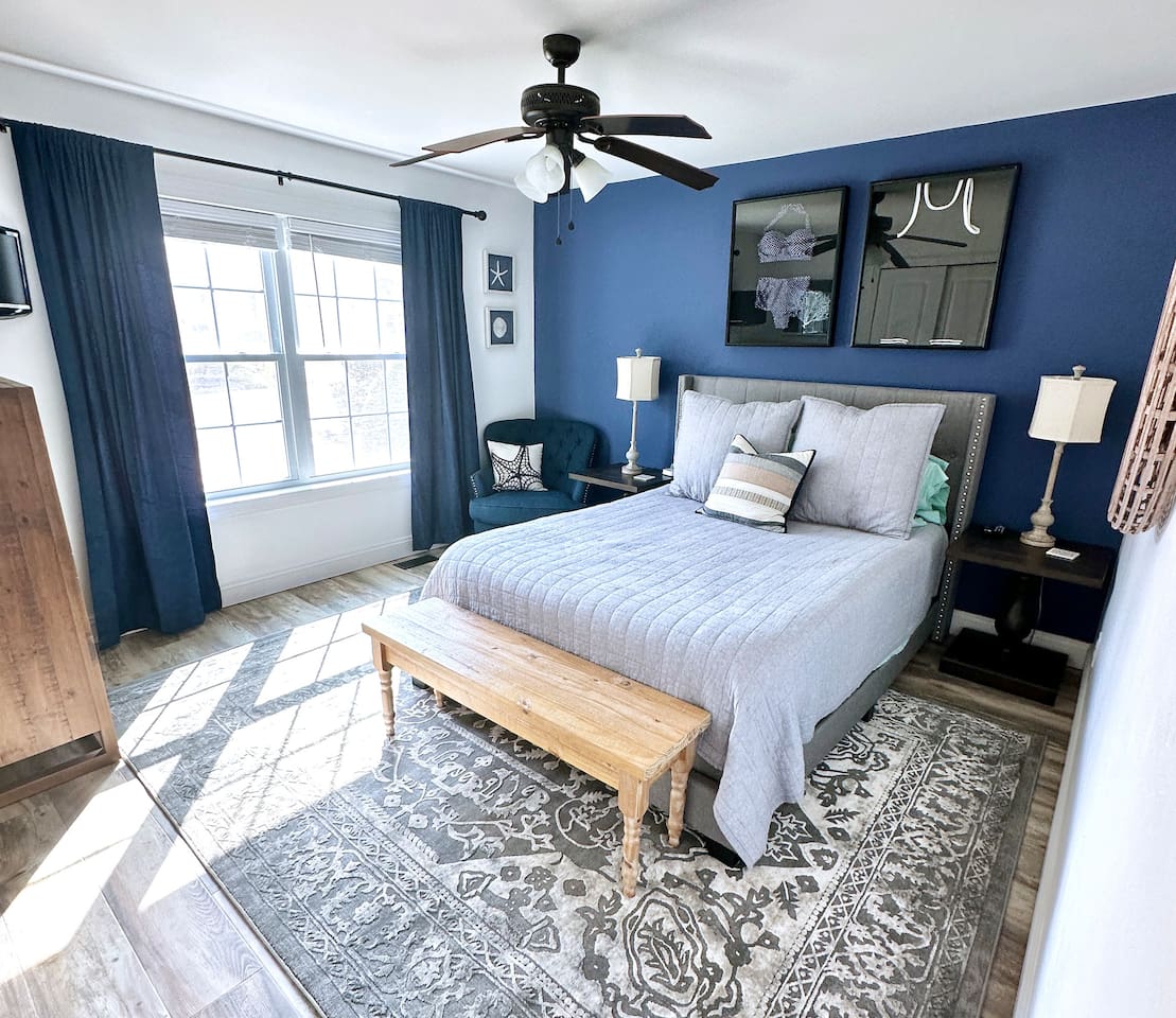 Relax in this sophisticated bedroom featuring a bold navy accent wall and a cozy upholstered bed. Guests can enjoy the soft natural light from the large window, a quiet reading nook in the corner, and a stylish wooden bench for added convenience.
