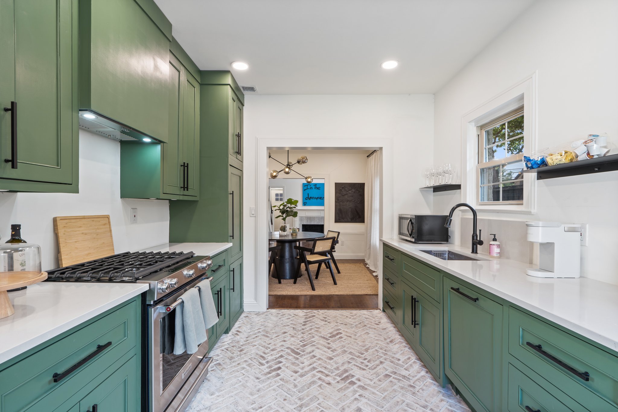 Kitchen with sage green cabinetry, gas range, and open view to dining area. Fully stocked with cookware, dishes, silverware, cooking basics, wine glasses, and Keurig coffee maker. Dishwasher included.