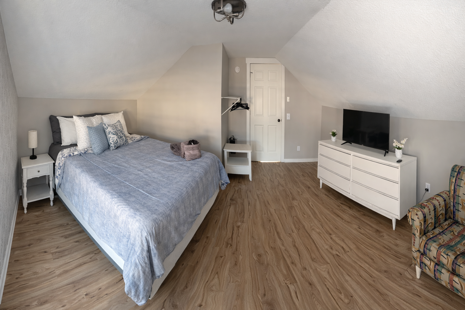 “Spacious upstairs bedroom with a comfortable queen bed, dresser with Smart TV, seating area, and open closet space. The warm wood floors and vaulted ceilings add charm and character.”