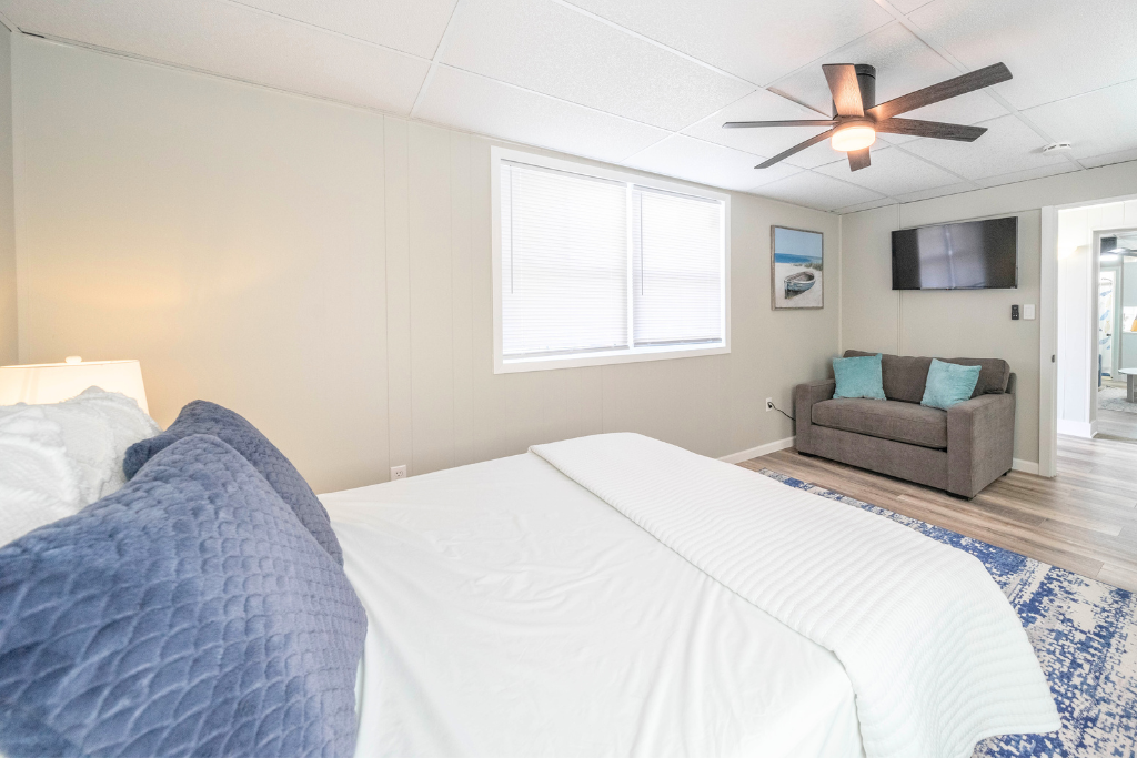 Spacious bedroom with a comfortable bed, soft linens, and calming coastal tones. Includes a sofa bed, wall-mounted TV, and ceiling fan, creating a relaxed space for rest, lounging, or winding down after a day at the beach.
