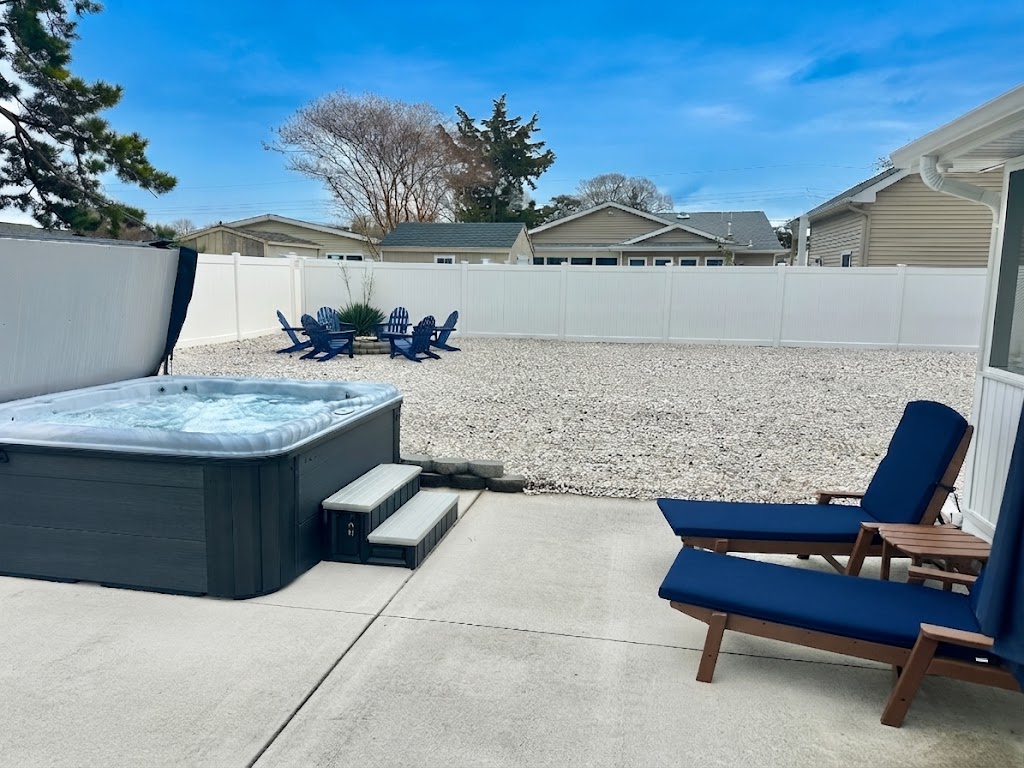 Unwind in your private outdoor retreat featuring a bubbling hot tub and comfortable lounge chairs with navy cushions. The spacious, fenced-in yard also includes a gravel area with Adirondack chairs gathered around a fire pit, perfect for starlit evenings.