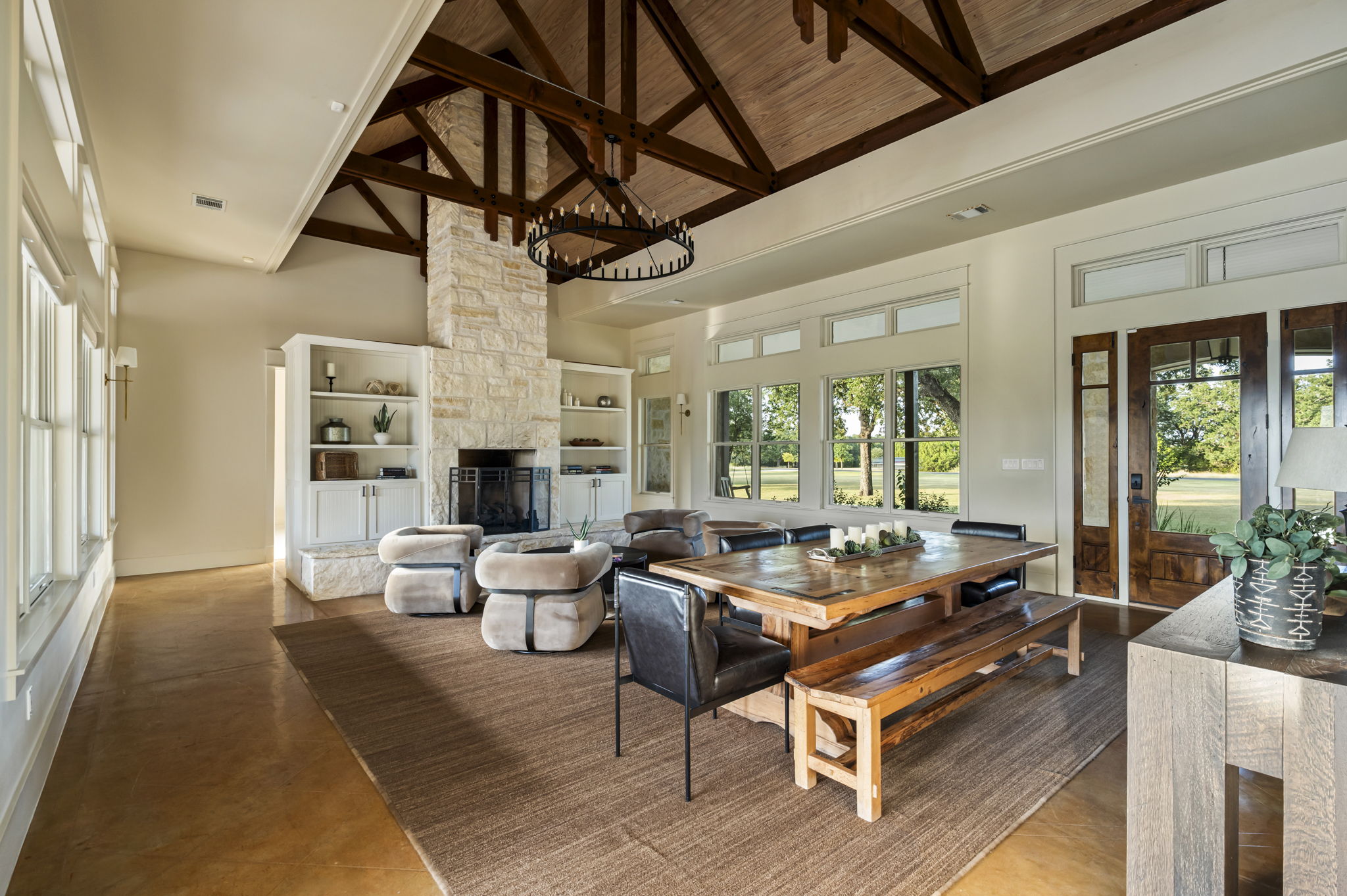 A spacious and inviting living area featuring high ceilings with exposed beams, a large dining table with benches, a cozy fireplace, and ample natural light.