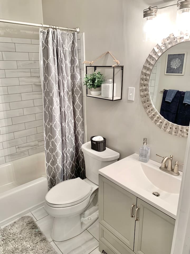 This modern full bath features a clean white vanity and a subway-tiled tub/shower combo. Coastal decor, like the round textured mirror and framed coral art, creates a bright, refreshing space for your daily routine.