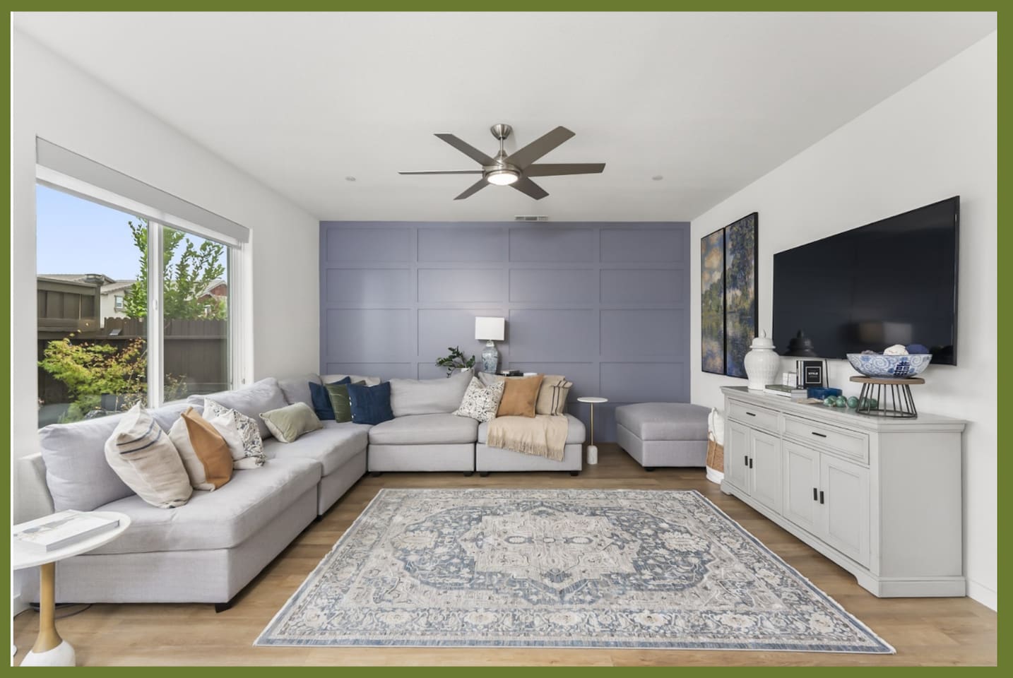 Bright living room with sectional seating, wall-mounted TV, ceiling fan, and a large window overlooking the backyard.
