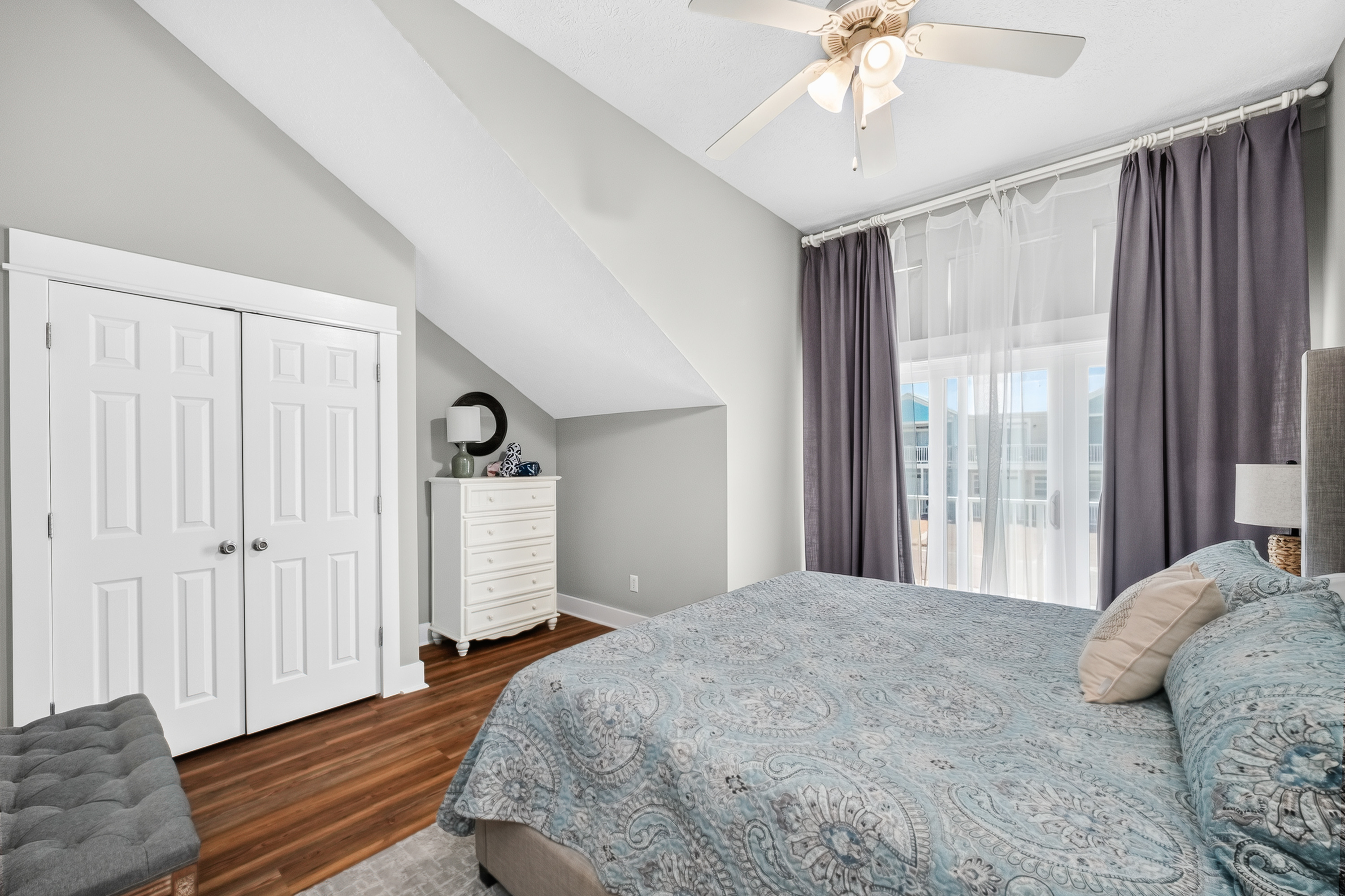 Comfortable king-size bed, plush linens, ceiling fan, spacious dresser, closet storage, bedside lamp, and a bright outdoor balcony.