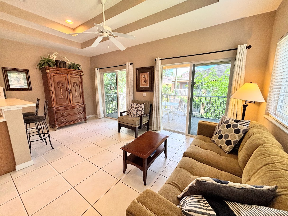 Comfortable living area with high ceilings, tile flooring, cozy seating, and two sets of sliding glass doors that bring in natural light and lead out to the private screened balcony.