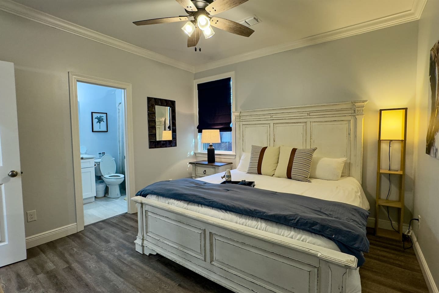 A bright, inviting bedroom with a classic white bed frame, layered neutral bedding, warm accent lighting, and direct access to a private ensuite bathroom for added comfort.