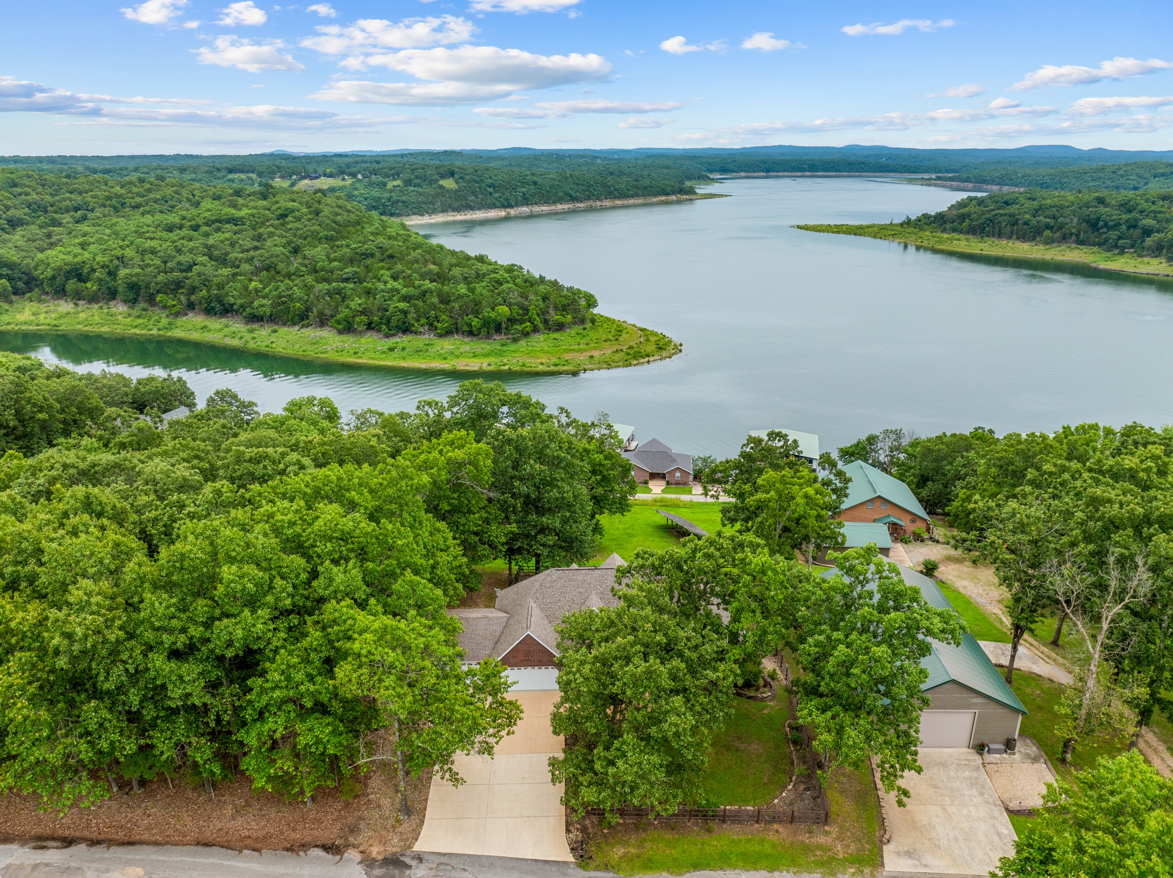 Aerial of the house above the lake cove, with a dock in the distance. Lake views stretch out beyond the trees.