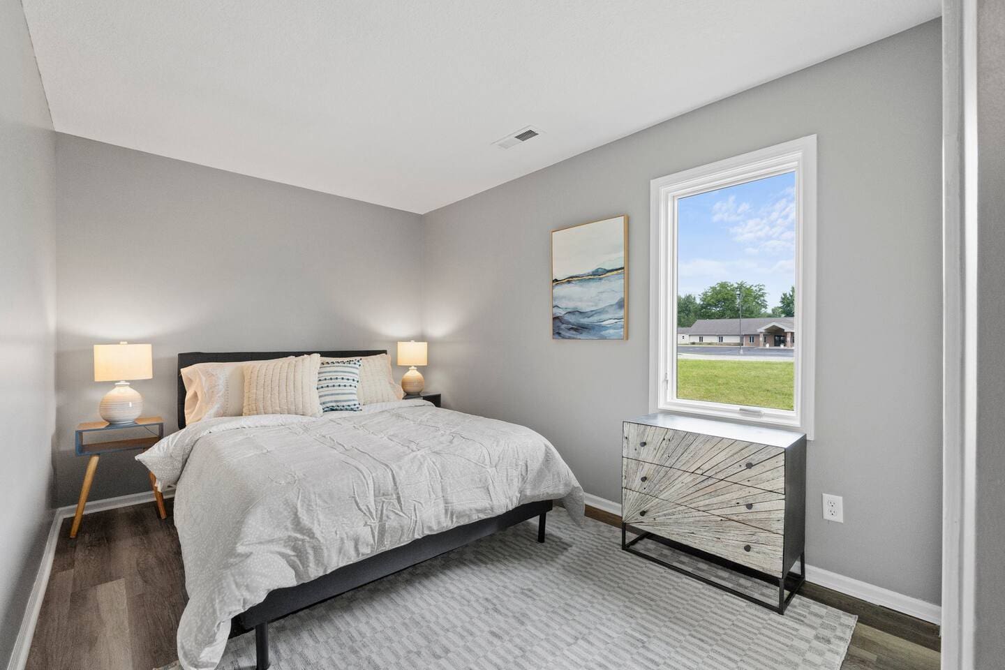 Charming queen-sized bed complemented by a stylish side table and lamps, with a window offering a beautiful view of the outside, creating a serene and cozy atmosphere.