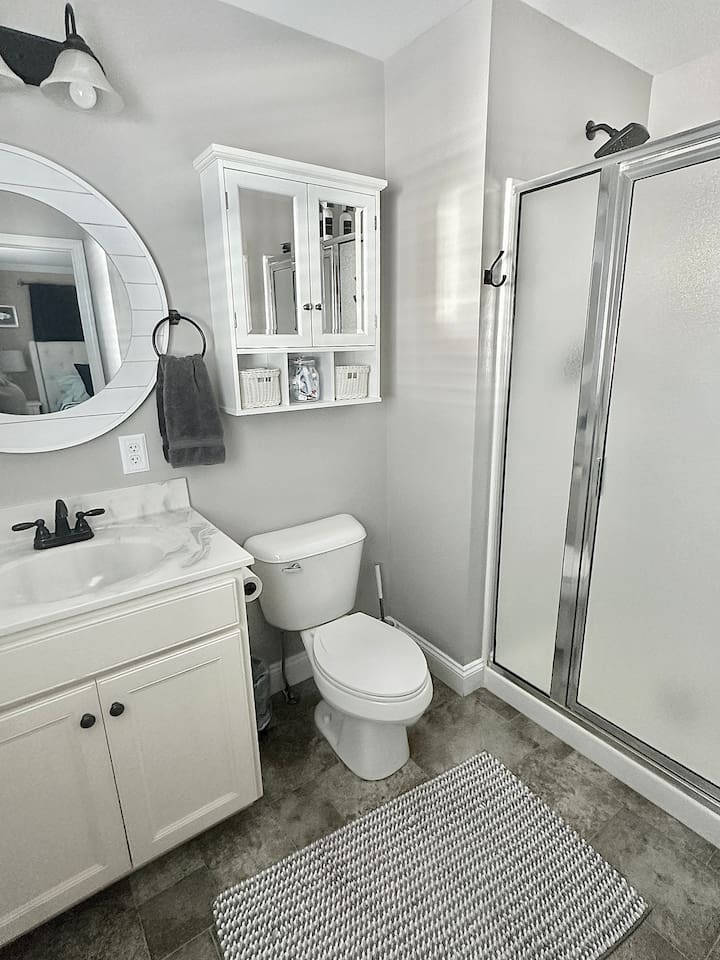 This bathroom features a white vanity with a marble-style top and dark hardware. The space includes a standard toilet, a walk-in shower with frosted glass doors, and grey tile flooring accented by a textured bathmat.