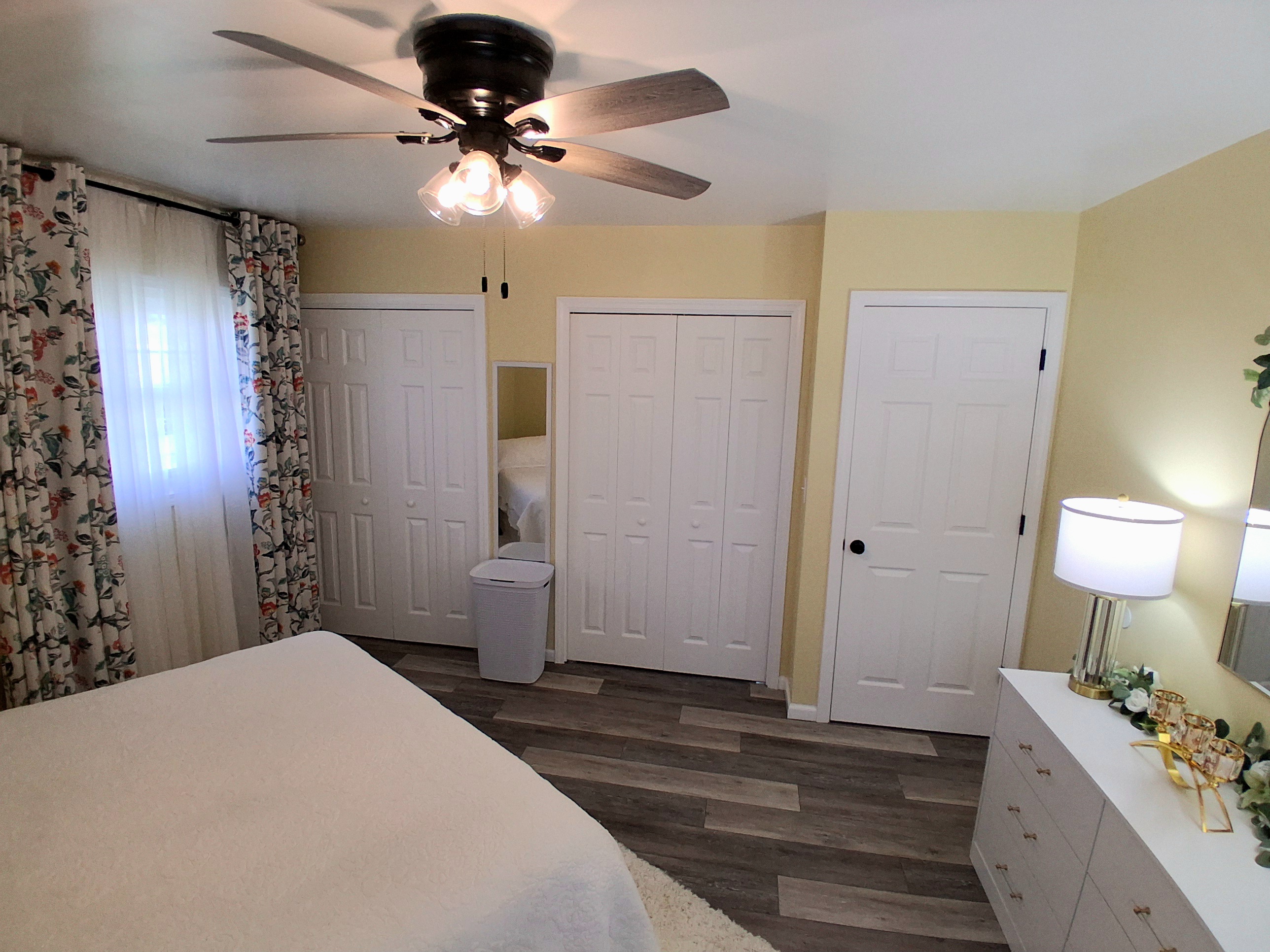 This sunny master suite feels spacious and uplifting, with soft yellow walls and floral curtains that brighten the room. Two roomy his-and-her closets offer generous storage, and natural light creates a peaceful, cheerful vibe all day.