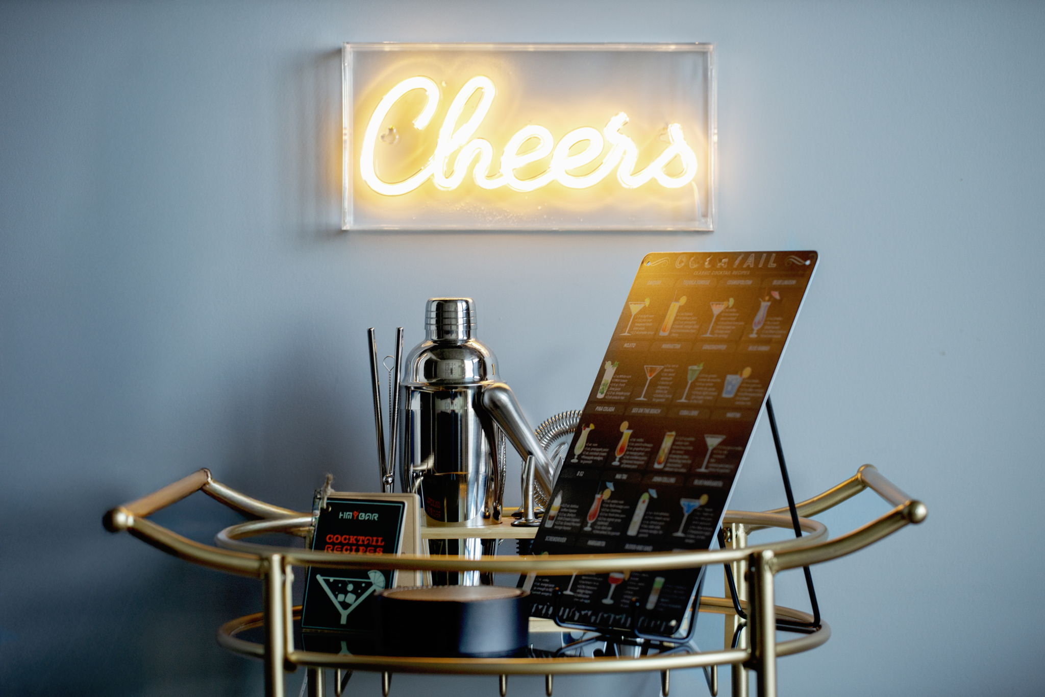 A gold bar cart styled with a cocktail shaker, mixing tools, and a guide to various drinks. A neon "Cheers" sign hangs on the gray wall above, creating a fun and inviting atmosphere.