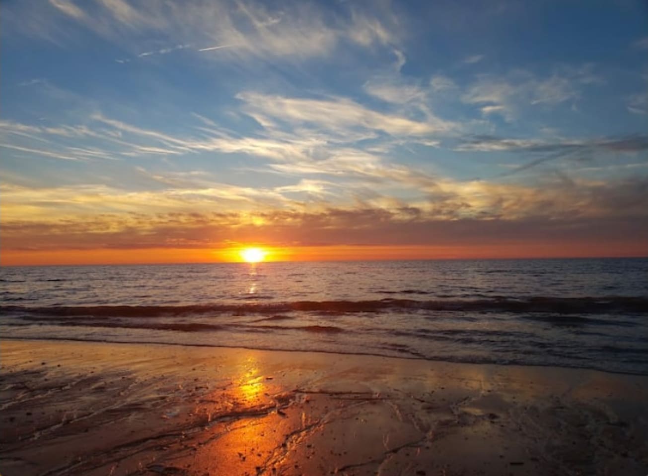 Take in the stunning horizon where the setting sun meets the ocean waves, casting a warm golden light across the wet sand. The wispy clouds add a dramatic touch to this tranquil coastal scene, located just a short walk from your front door.