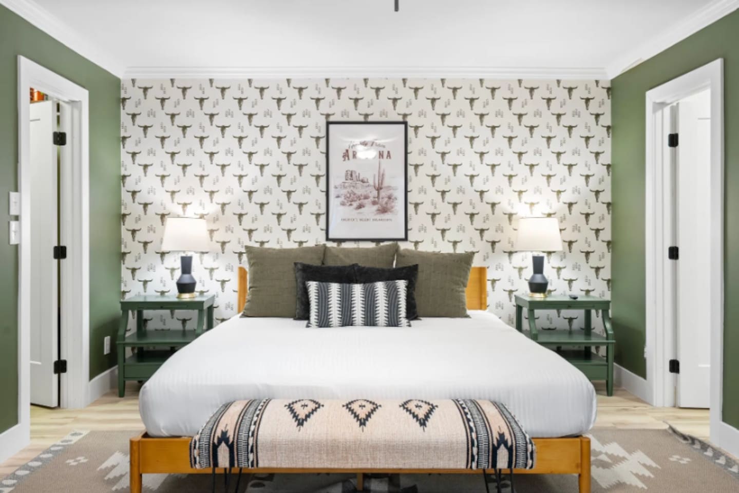 A king bed dressed in crisp white linens sits under a striking desert-print wallpaper, with matching nightstands and warm lighting on both sides. Every detail here was chosen to feel curated, not cluttered.
