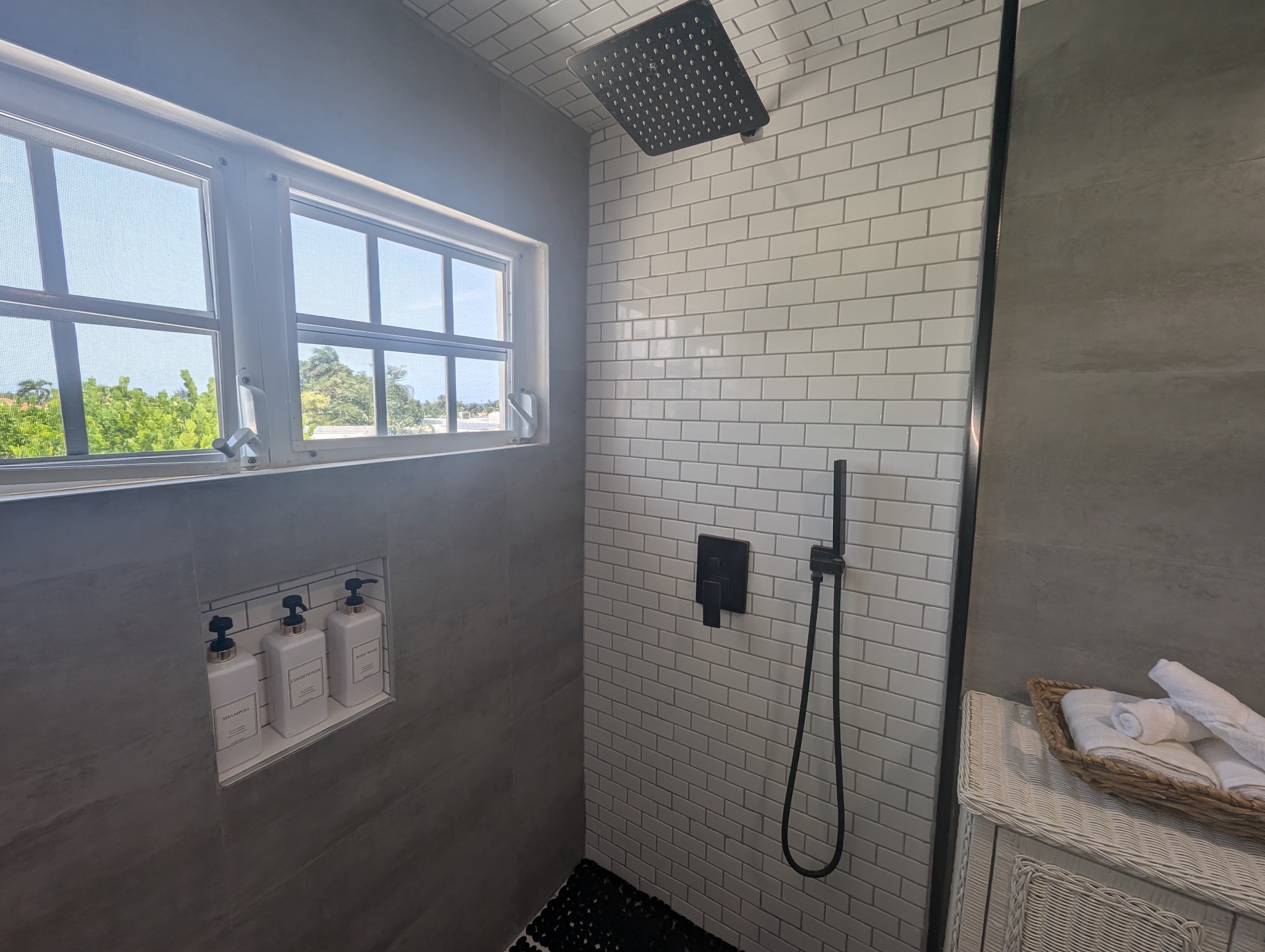 Walk-in shower at Villa Coral showcasing a matte black square rainfall showerhead, a matching handheld sprayer wand, white subway tiles, and built-in toiletry dispensers under bright windows.
