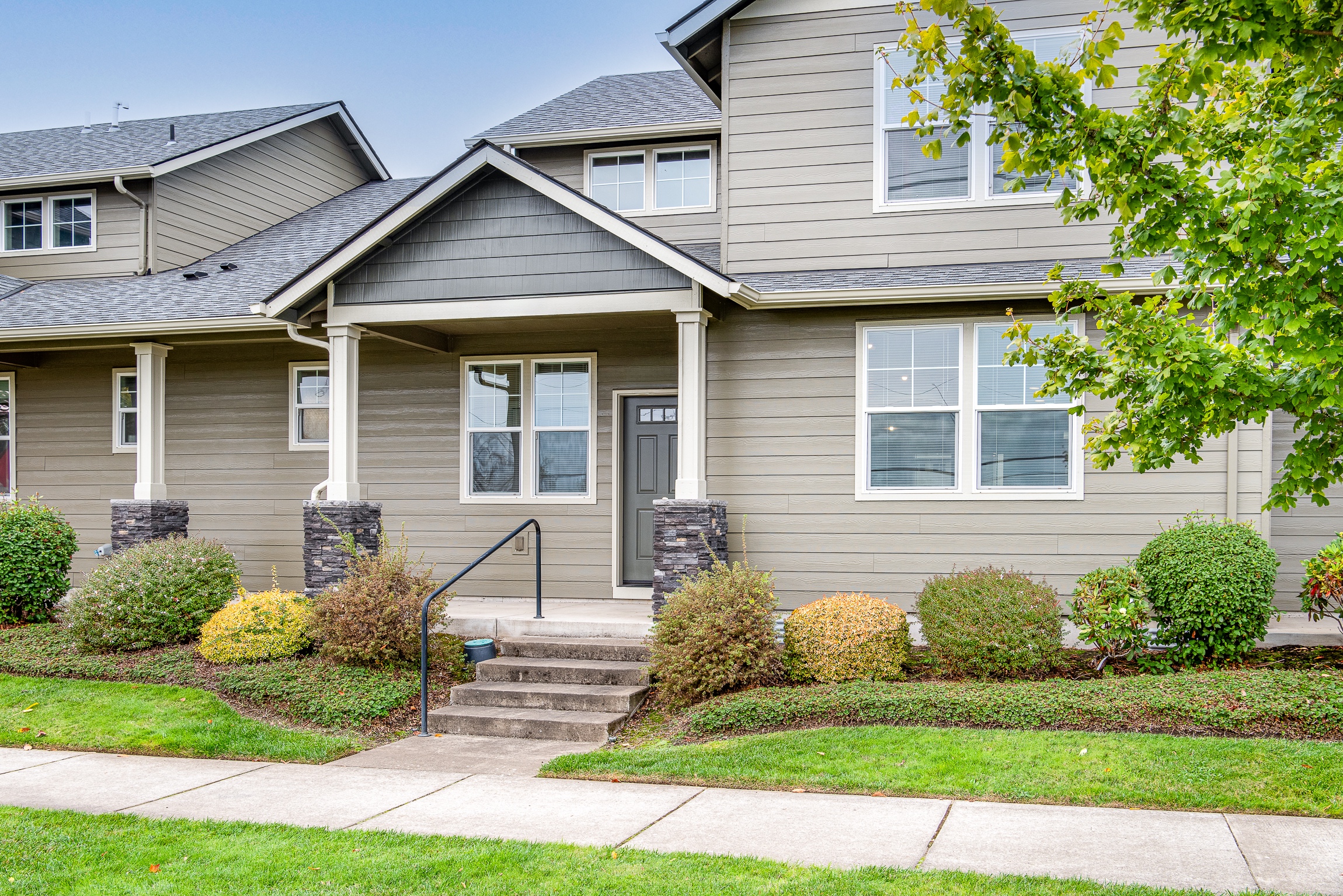 A modern two-story house with gray siding, a covered front porch supported by white columns, a slate stone base, decorative shrubs, and well-maintained grass, situated next to a sidewalk.