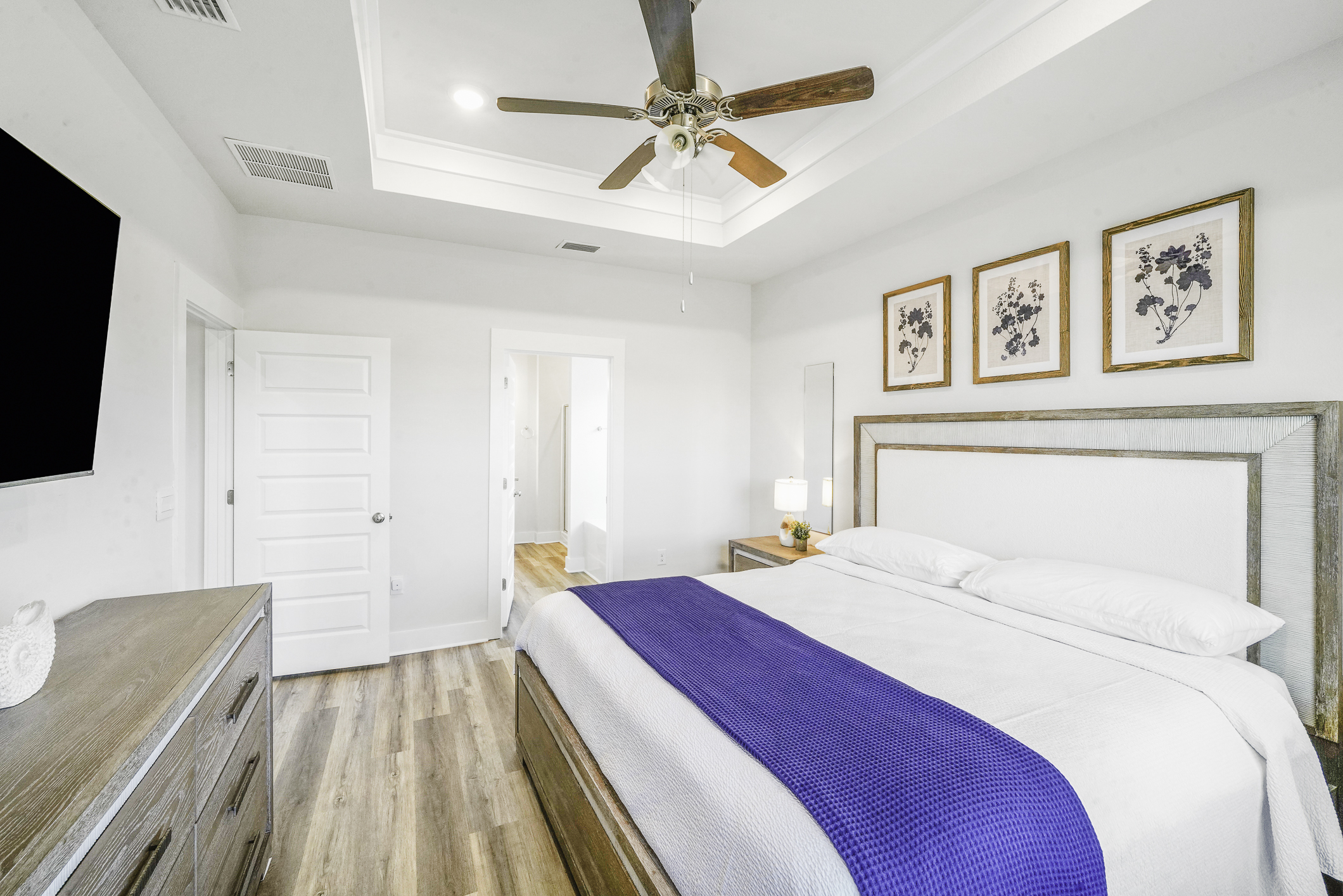 Primary Suite: Modern king bedroom featuring an en suite bathroom, plush linens, ceiling fan, elegant bedside lamps, dresser, and Smart TV.