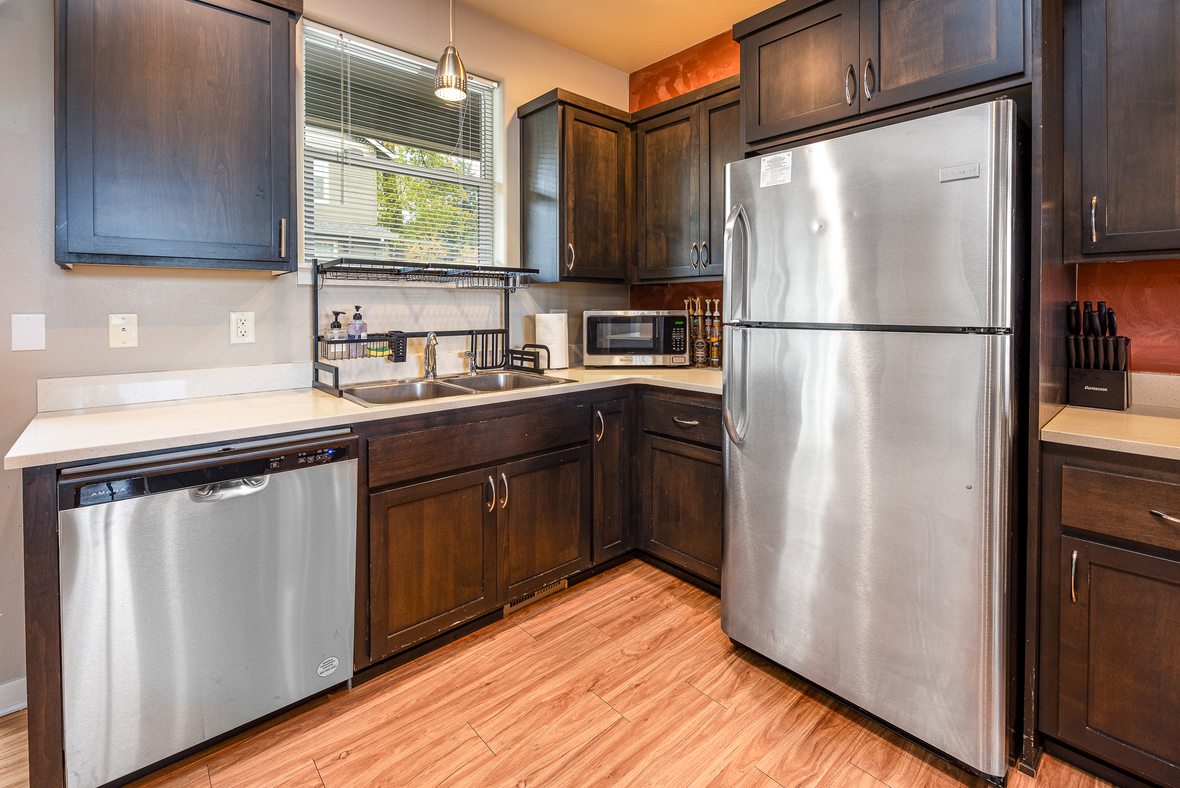 A modern kitchen featuring dark wood cabinets, stainless steel appliances including a refrigerator and dishwasher, a double sink with a chrome faucet, a countertop with decorative dishware, and a microwave, all set against a warm-colored wall and wooden floor.