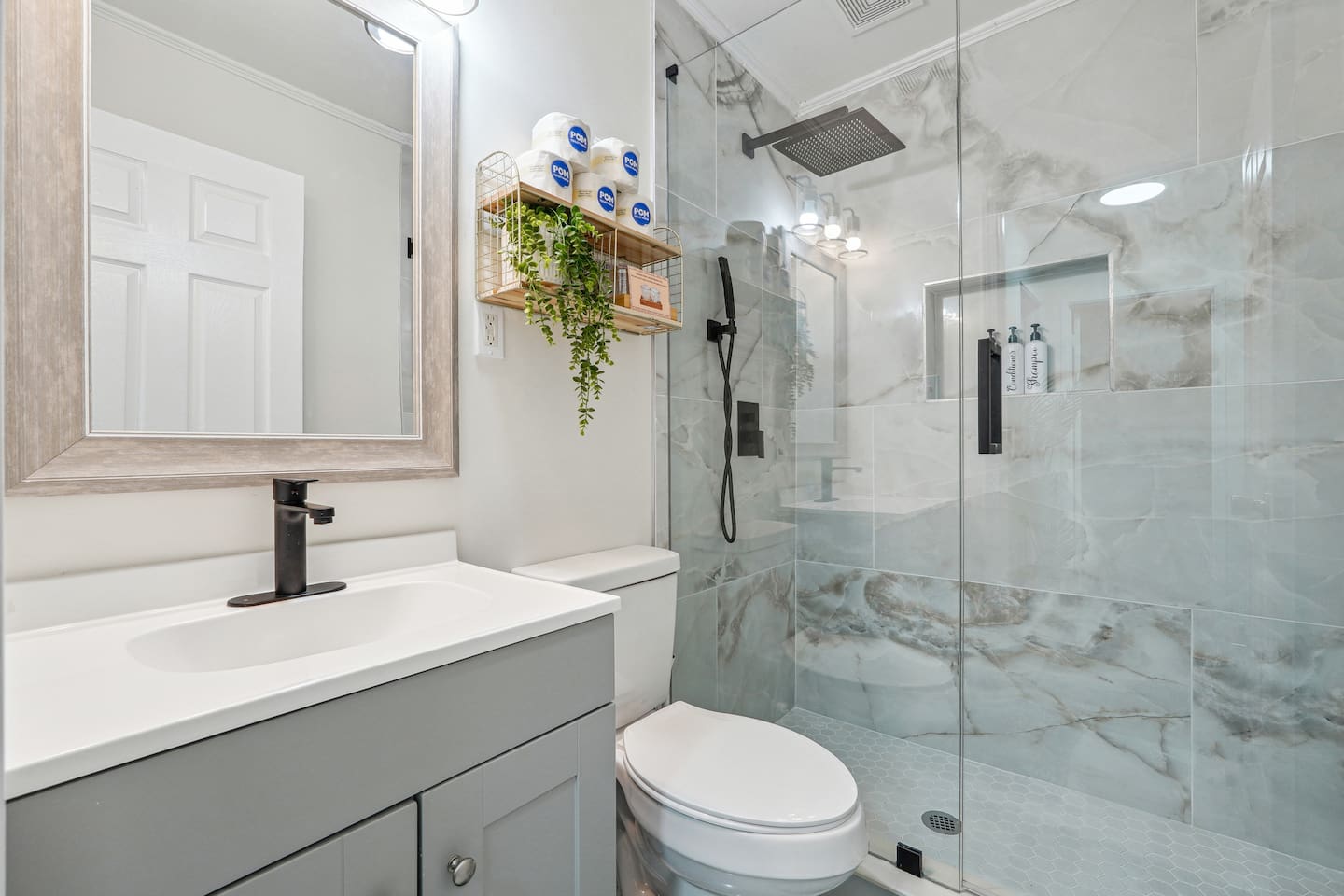 A clean and well-equipped bathroom featuring a mirror, sink, toilet with toilet paper provided, and a shower with both hot and cold water. Essentials like shampoo, conditioner, and body wash are also available for your convenience.
