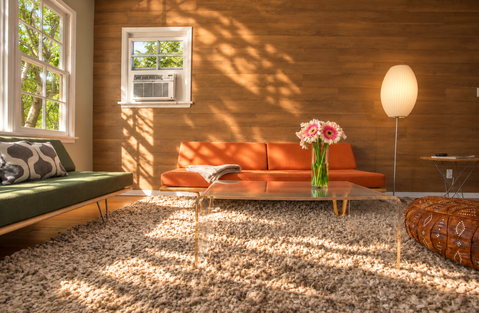 The sunlit living room combines an orange sofa and green seating with a clear acrylic coffee table, textured rug and leather pouf. Leaf-shaped shadows play across the wood feature wall, and a glowing floor lamp adds warmth beside the sofa.