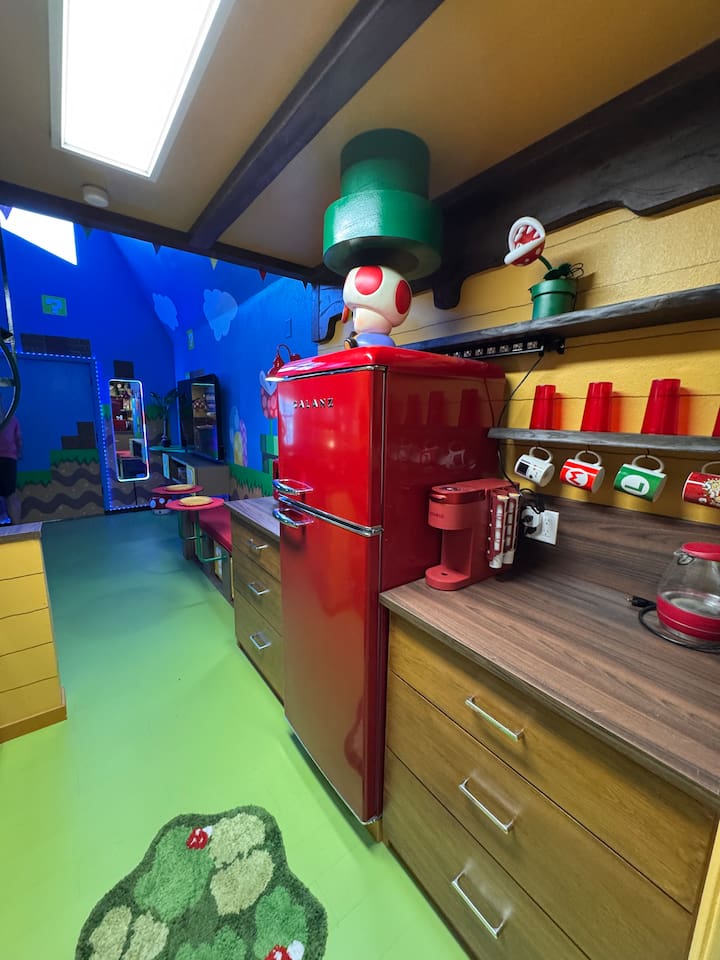 Whimsical full-yet-compact kitchen at Mario's Mini Mansion, Clermont, FL! Bright red retro-style fridge, green flooring, yellow walls with Mario shelves, and Toad-inspired decor make post-theme park meal prep an adventure for kids and adults alike.