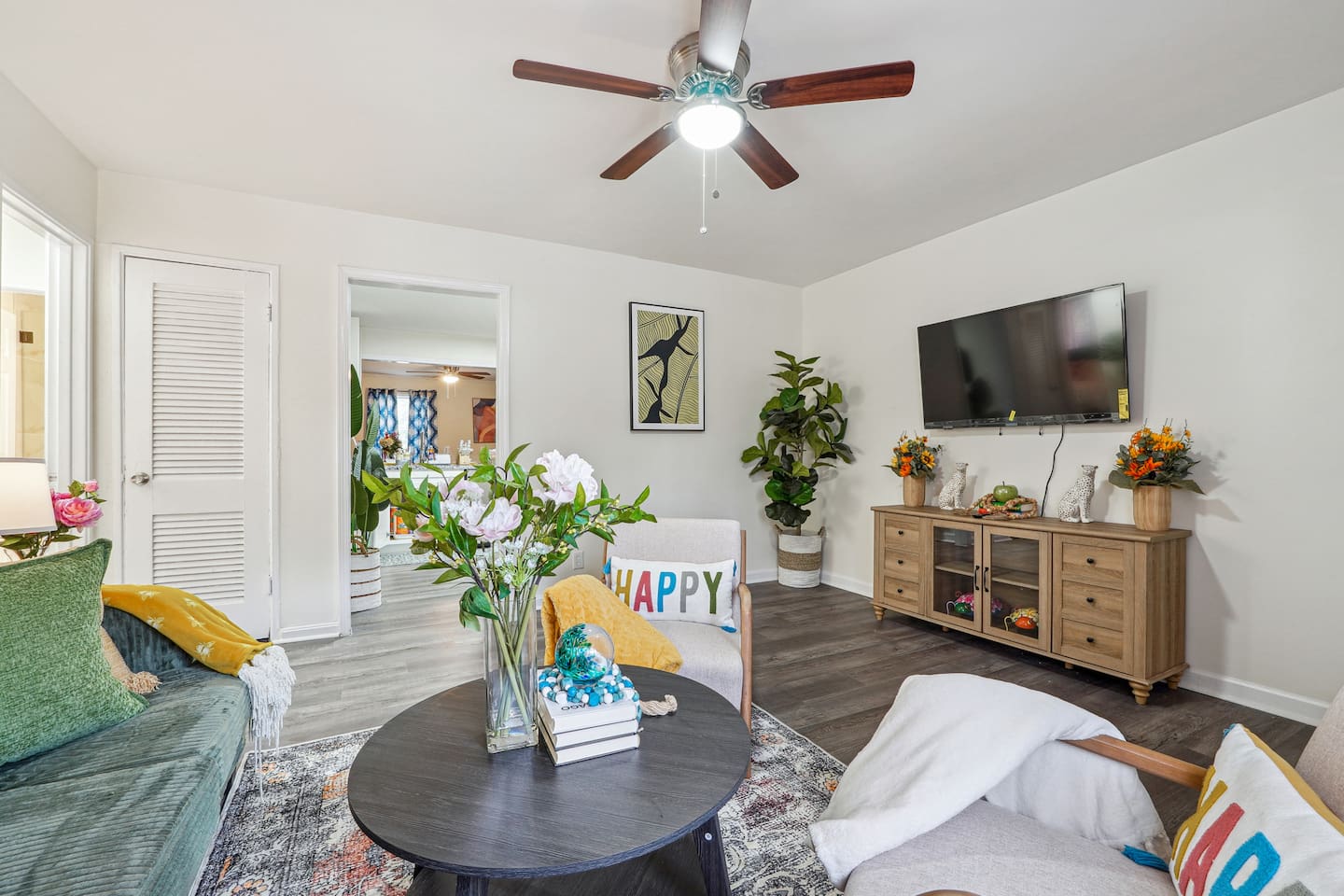 Enjoy a cozy living room featuring a long comfortable sofa, a TV for your entertainment, a ceiling fan, two chair, and a window with a lovely view outside — perfect for relaxing and unwinding.
