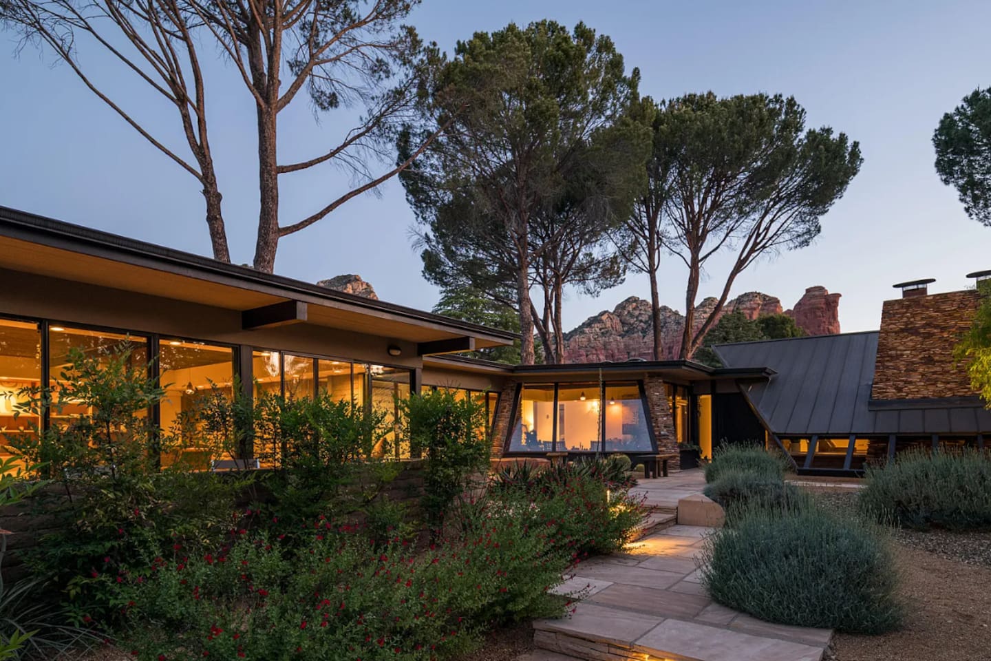 Stunning mid-century modern home at dusk, showcasing dramatic rooflines, floor-to-ceiling windows, and captivating red rock views beyond beautifully landscaped gardens.