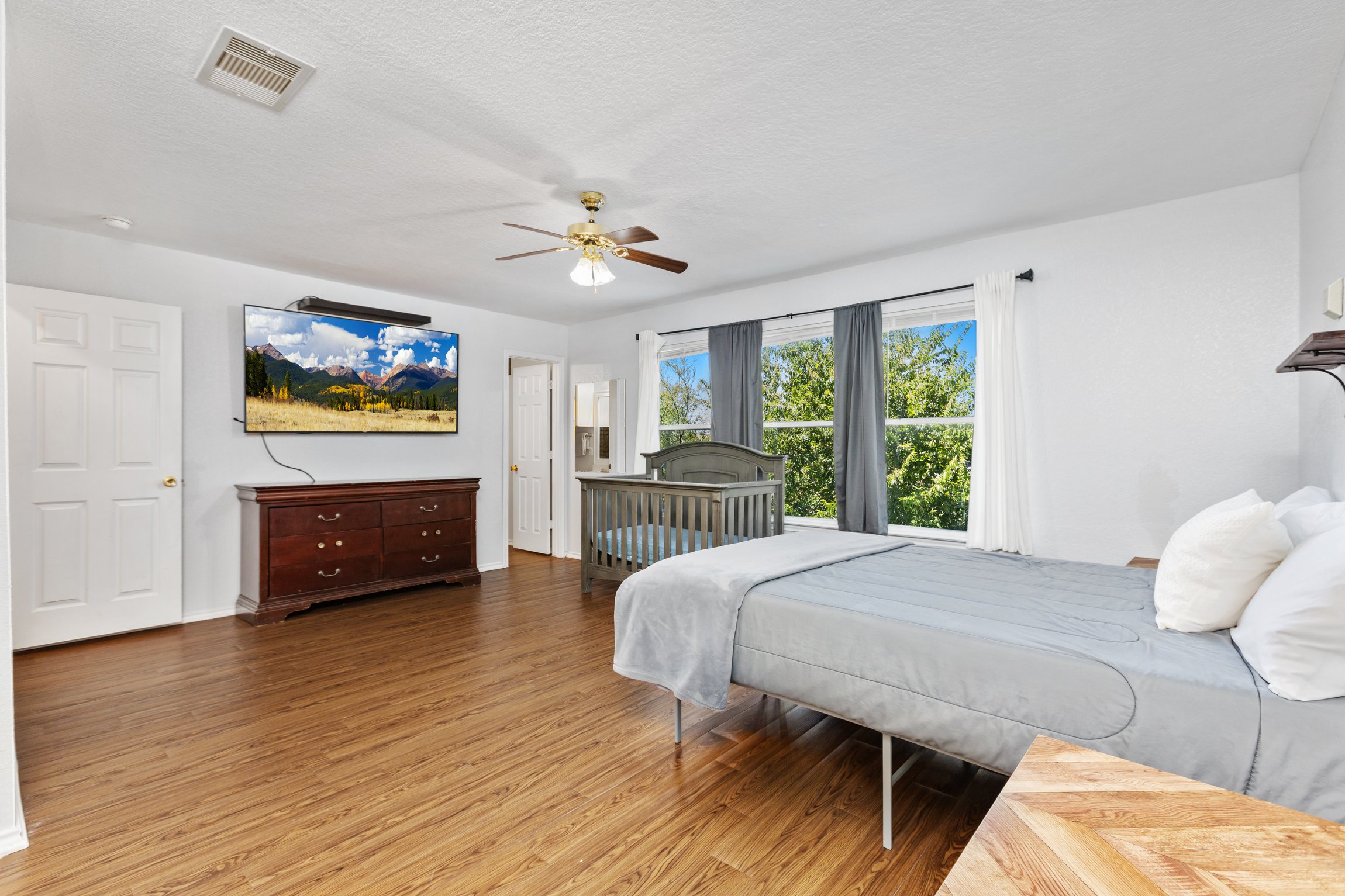 King bedroom with wall-mounted Smart TV, ceiling fan, natural light, gray bedding, and room-darkening shades. Clothing storage and extra pillows included.
