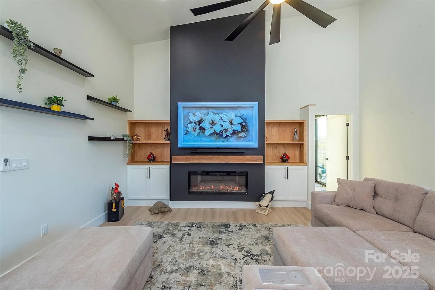 Bright and spacious open-concept living area featuring a modern electric fireplace, custom built-in shelving, cozy sectional seating, and soaring ceilings with a contemporary ceiling fan.