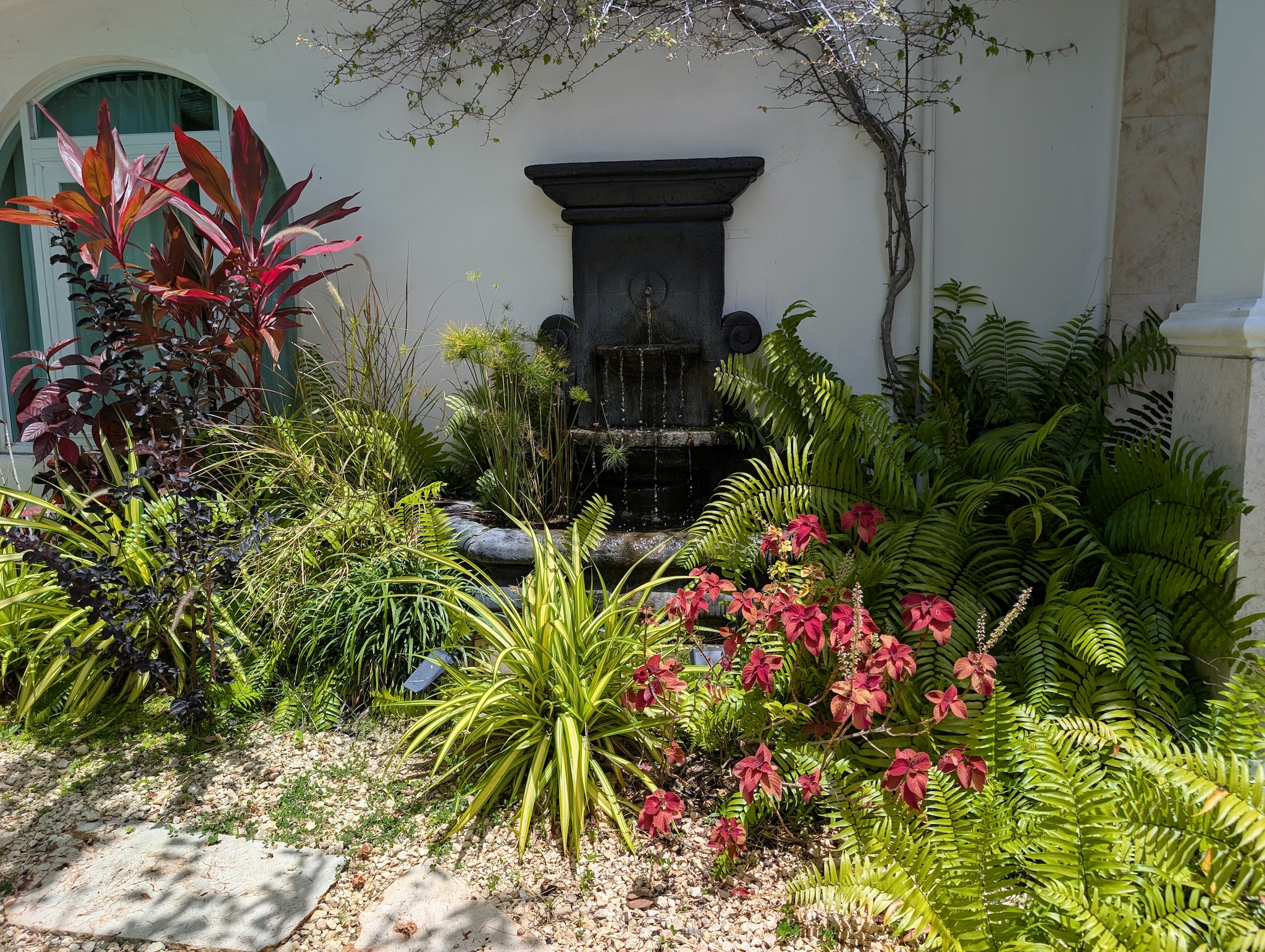 Stone tiered wall fountain surrounded by lush tropical landscaping, green ferns, and colorful plants at the entryway of Villa Coral in Dorado, Puerto Rico.
