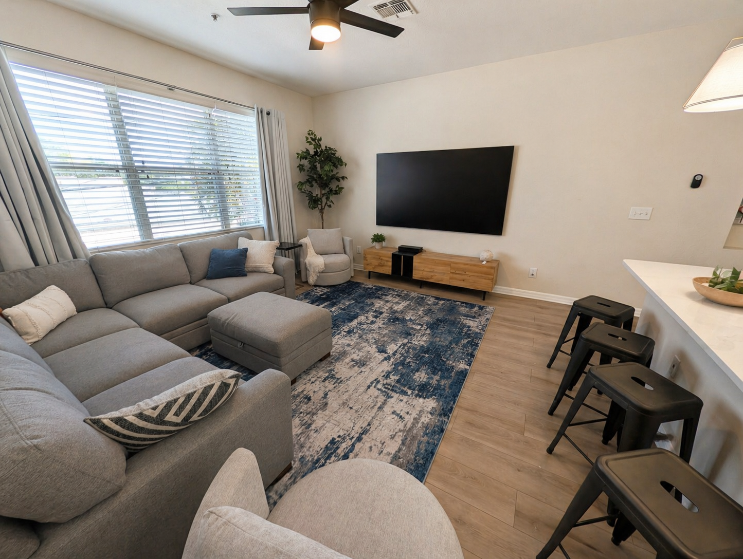 Bright, open living room with a large sectional sofa and ottoman—perfect for relaxing or gathering. Enjoy natural light from oversized windows, a big-screen TV, and easy access to the kitchen with bar seating for casual dining.