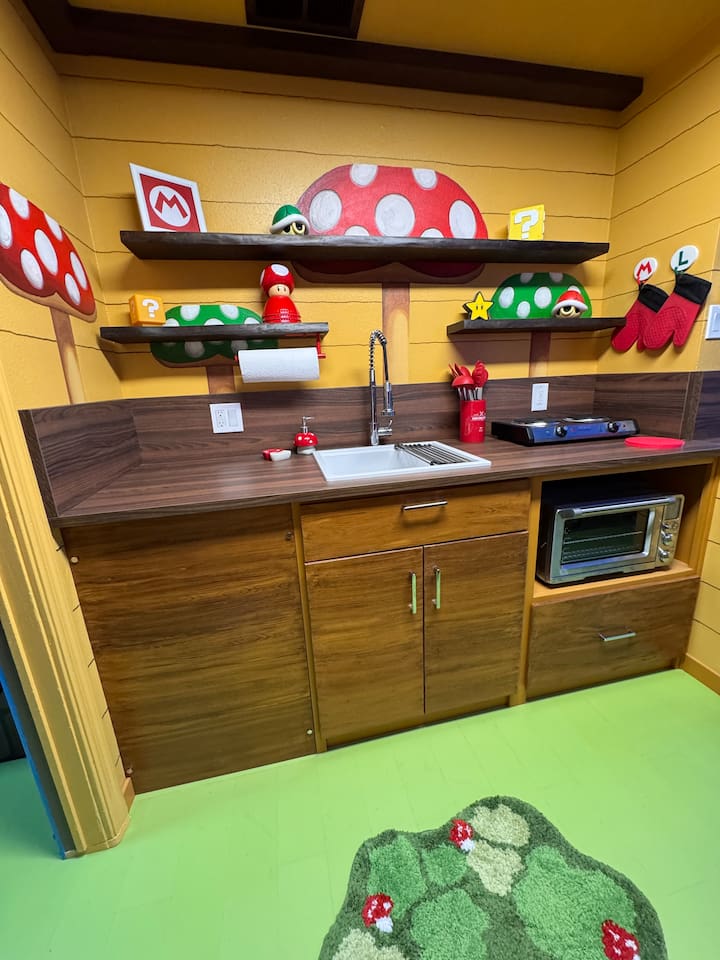 Fully equipped Nintendo-inspired kitchen! Warm wood cabinets, dark countertops, built-in microwave, sink, and vibrant Super Mario mushroom shelves stocked with fun details—perfect for family meals at Mario's Mini Mansion.