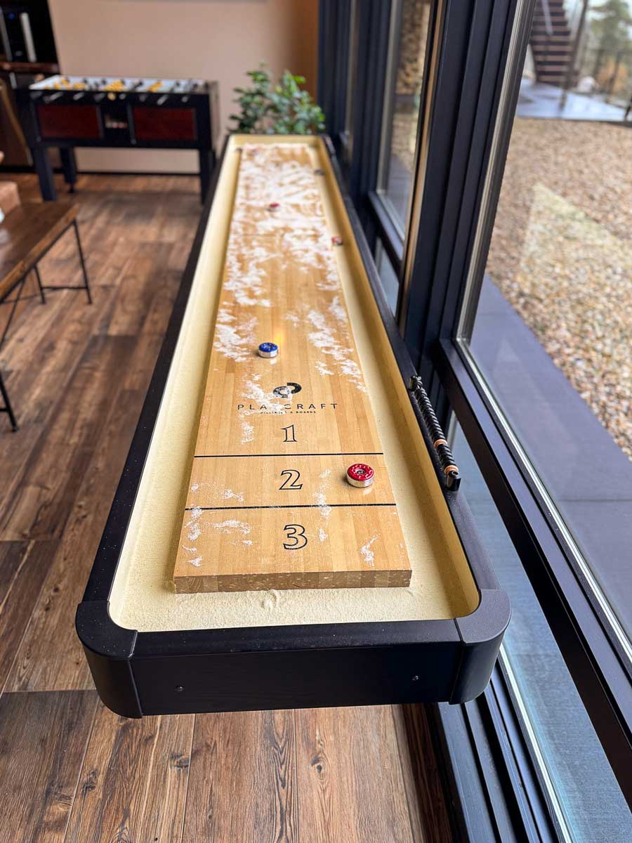 The Tavern Room at Sunset Falls &mdash; shuffleboard table along floor-to-ceiling glass with mountain view beyond