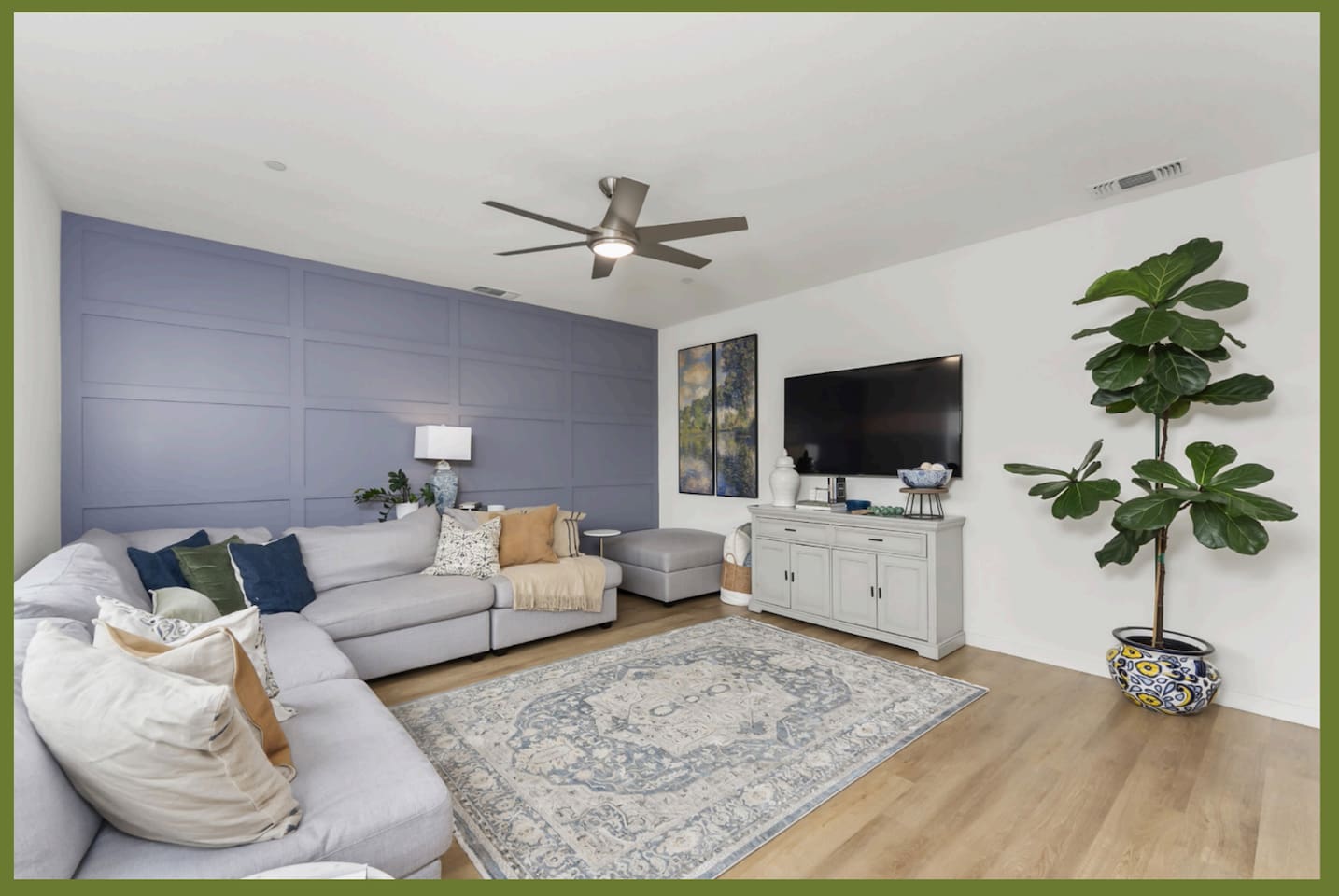 Bright living room with sectional seating, wall-mounted TV, ceiling fan, and a large window overlooking the backyard.