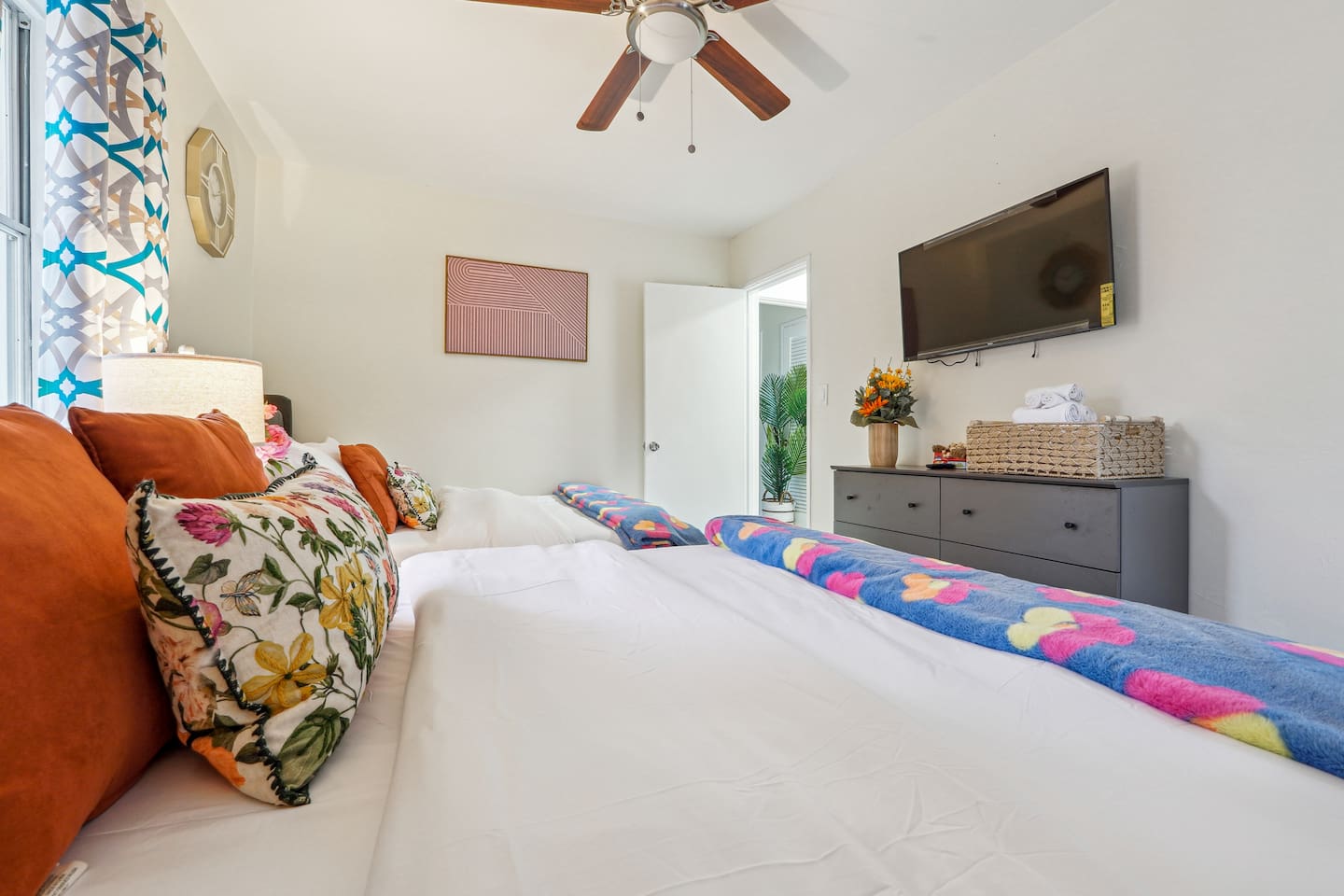 Step into a comfortable bedroom featuring two Queen beds, a ceiling fan, a TV, a clock, and a basket of fresh towels, providing everything you need for a relaxing and restful stay.
