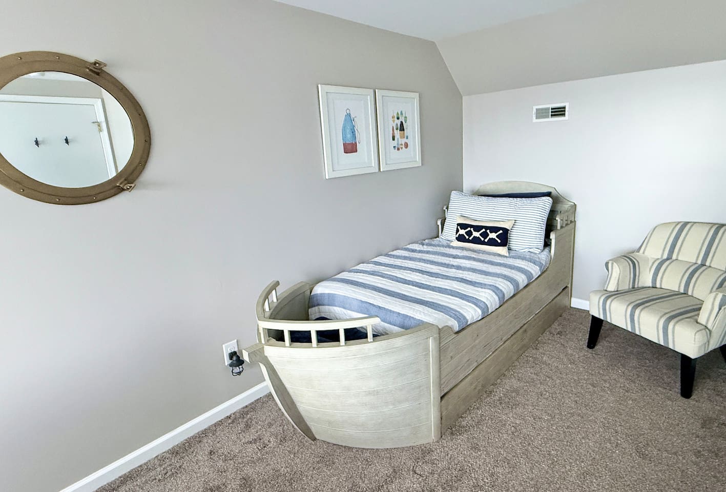 The kids will love the nautical-themed bedroom features a unique bed shaped like a boat, complemented by a round porthole-style mirror, striped accent chair, and framed coastal wall art to create a charming, sea-inspired retreat.