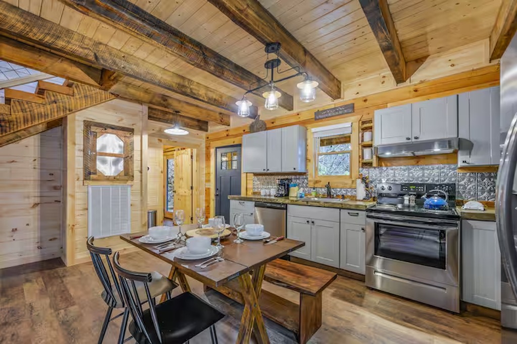 The kitchen and dining area at Bear Paws Cabin sit on the main level, wide open and family-friendly. Exposed timber ceiling beams, a farmhouse dining table, and modern stainless appliances &mdash; everything your family needs to settle in and feel at home