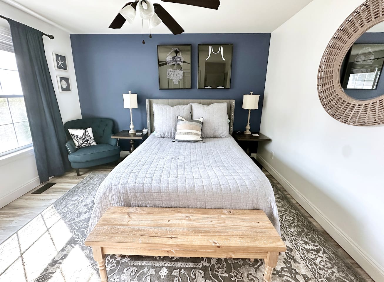 This modern bedroom features a blue accent wall and a neutral bed with a wooden end-of-bed bench. The space includes a teal armchair, unique wall art, and a large round mirror, all set over stylish hardwood-style flooring.