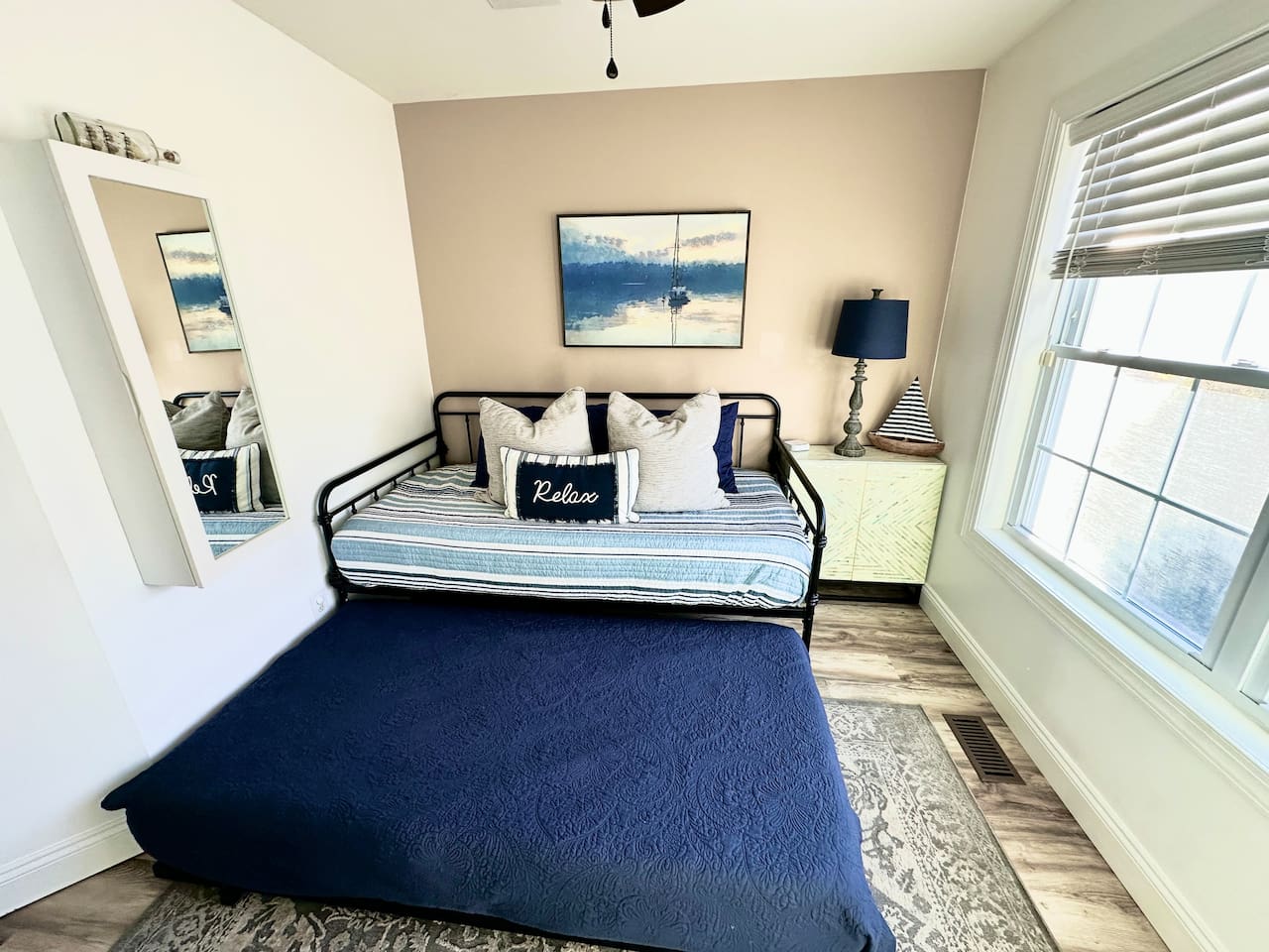 This functional room features a black metal daybed with a pull-out trundle bed dressed in a navy textured coverlet. The space includes nautical wall art, a light-green cabinet with a lamp, and a large window with blinds.