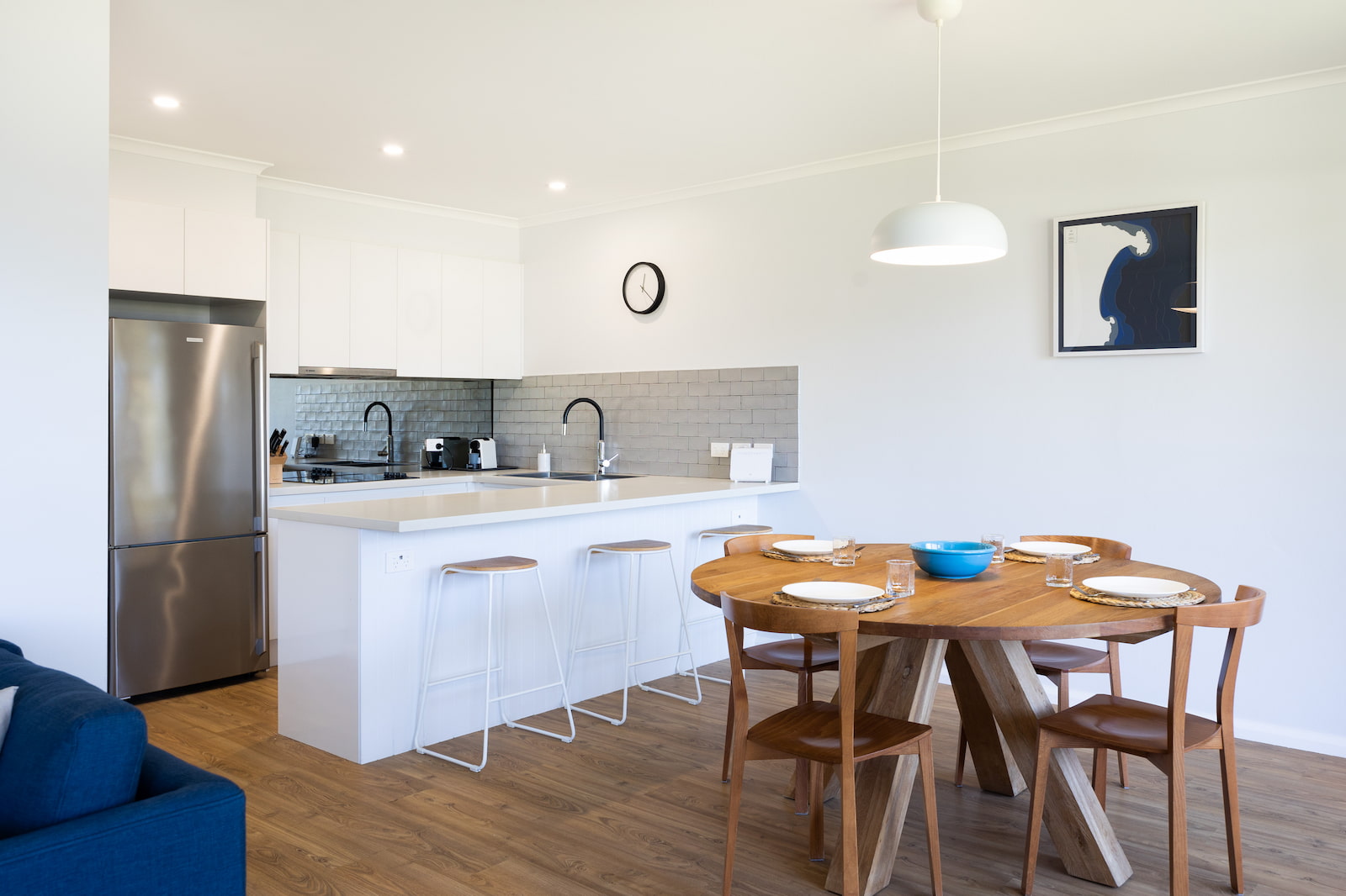 White kitchen counter with three white stools, grey tiles, stainless steel fridge, white cabinets and wooden dining table in foreground.