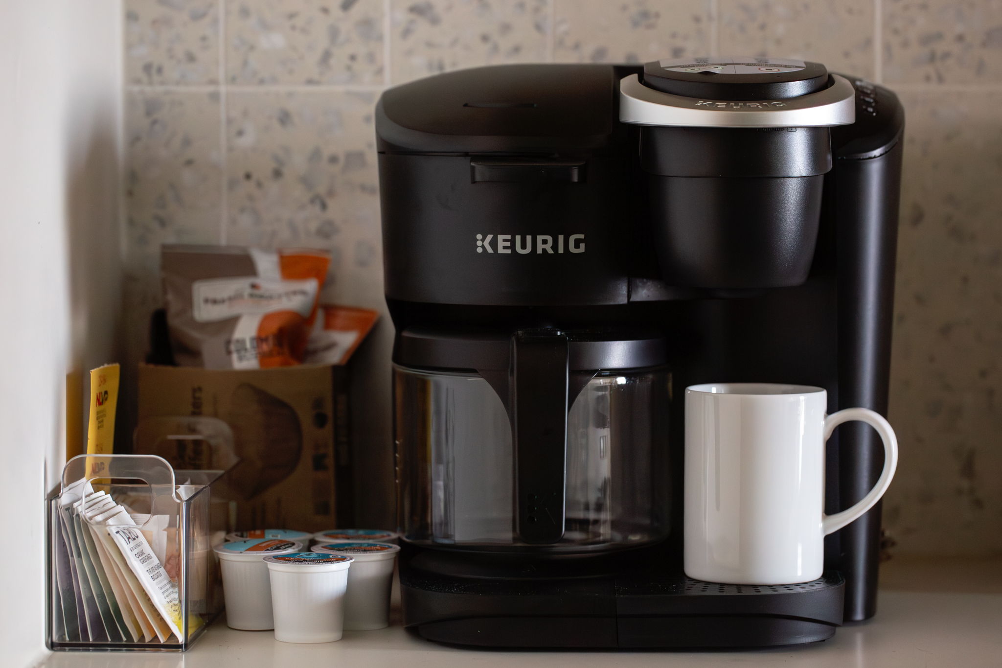 A dedicated coffee station featuring a black Keurig machine with a carafe and a single-serve K-Cup option. The setup includes an organizer with various sweeteners and stirrers, a small selection of K-Cups, and a crisp white mug, all placed on a countertop