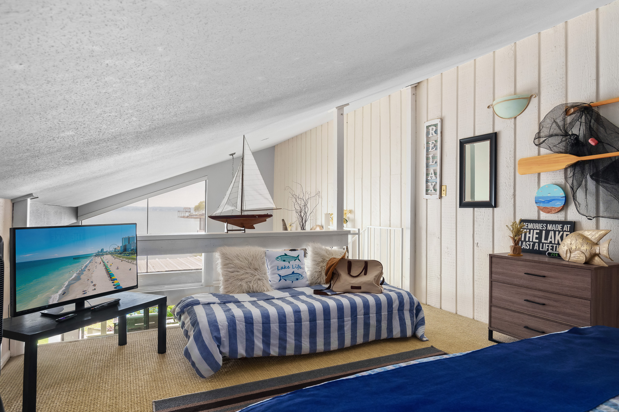 This cozy loft bedroom features a king bed and twin bed, Roku TV with cable, dresser, and fan—all tucked under charming sloped ceilings with lake views. Perfect for families or extra guests looking to unwind in comfort.