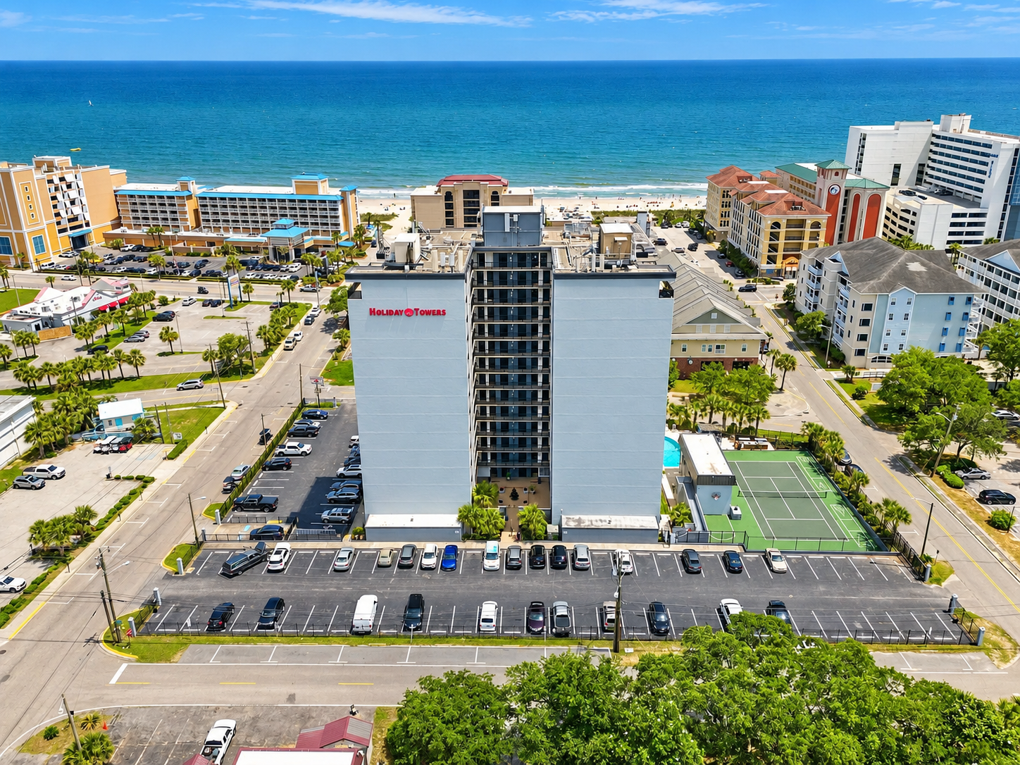 Aerial view of Holiday Towers with shared pool, hot tub, sports court, parking, and Myrtle Beach attractions nearby.