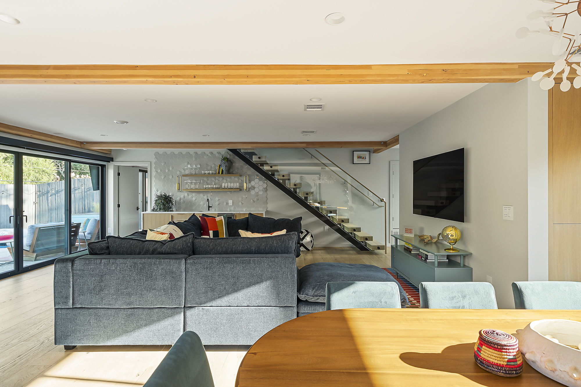 A spacious open-concept living area featuring exposed wooden beams, a large sectional sofa, a modern staircase with a glass railing, and sliding doors that open to an outdoor space.