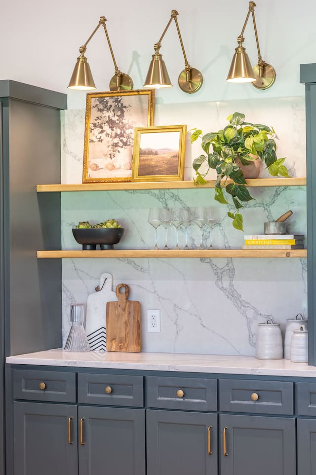 Styled open shelving detail featuring brass sconces, marble backsplash, curated décor, and glassware for a polished kitchen look.