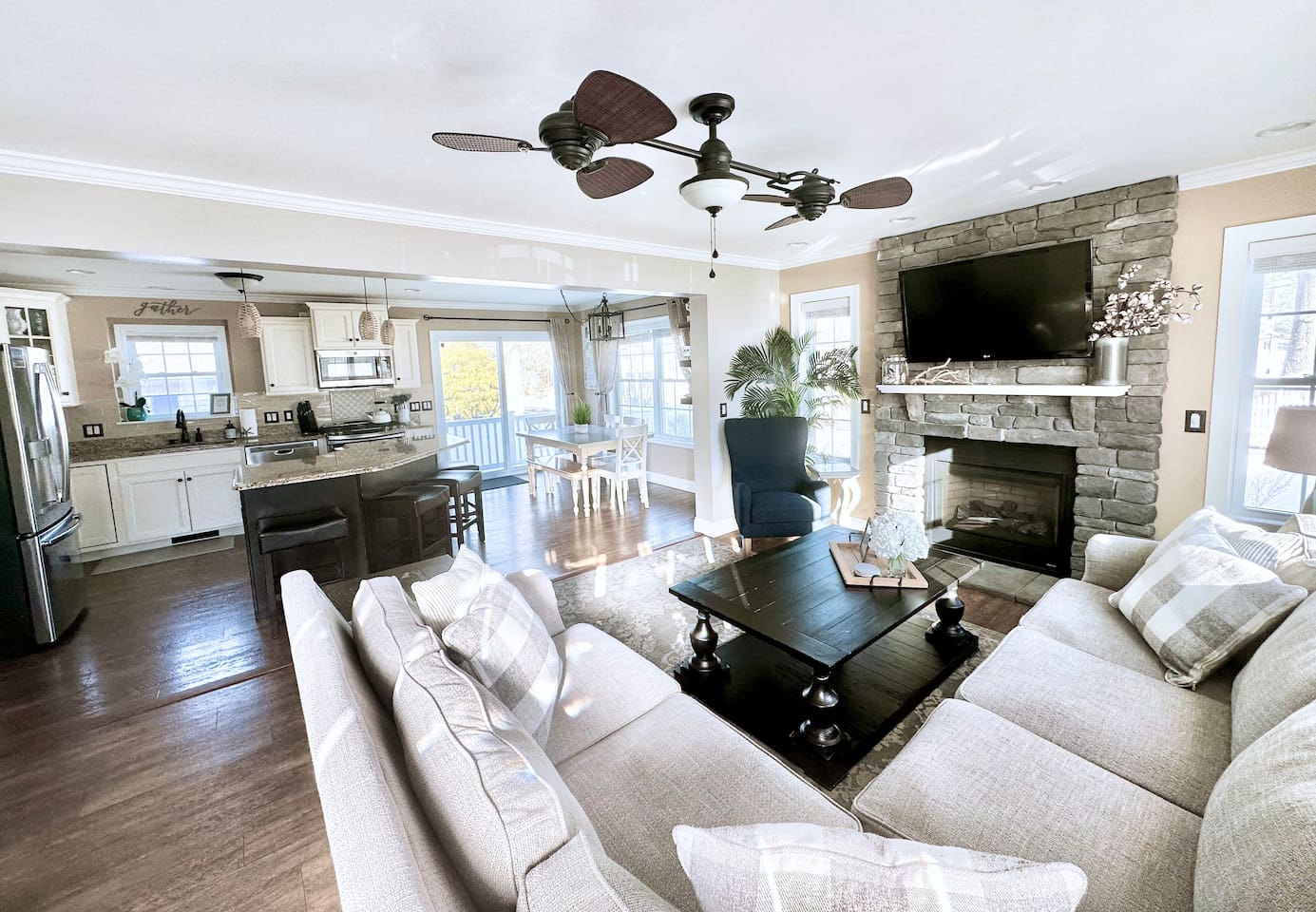This wide view showcases the seamless flow between the cozy living room, modern kitchen, and dining area. The open-concept design is anchored by a stone fireplace and a unique ceiling fan, creating a spacious and unified gathering spot.
